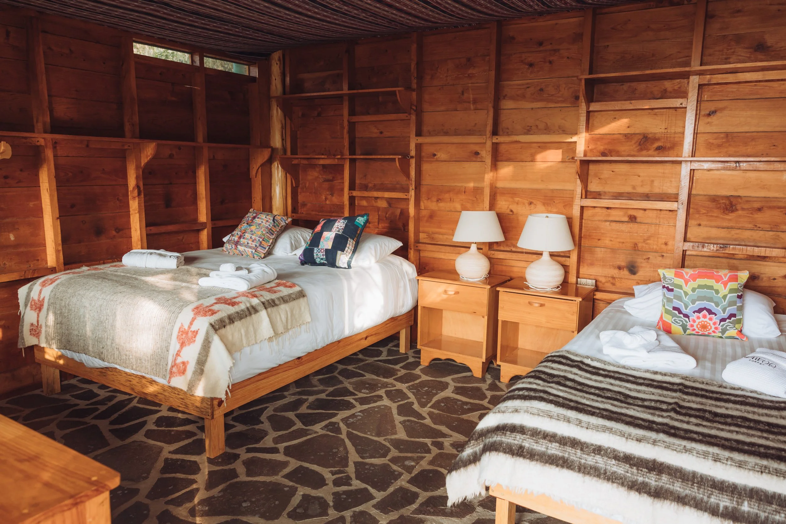 A rustic bedroom with wooden walls and two beds, each with colorful pillows and blankets. Two bedside tables with lamps are between the beds, set on a stone floor.