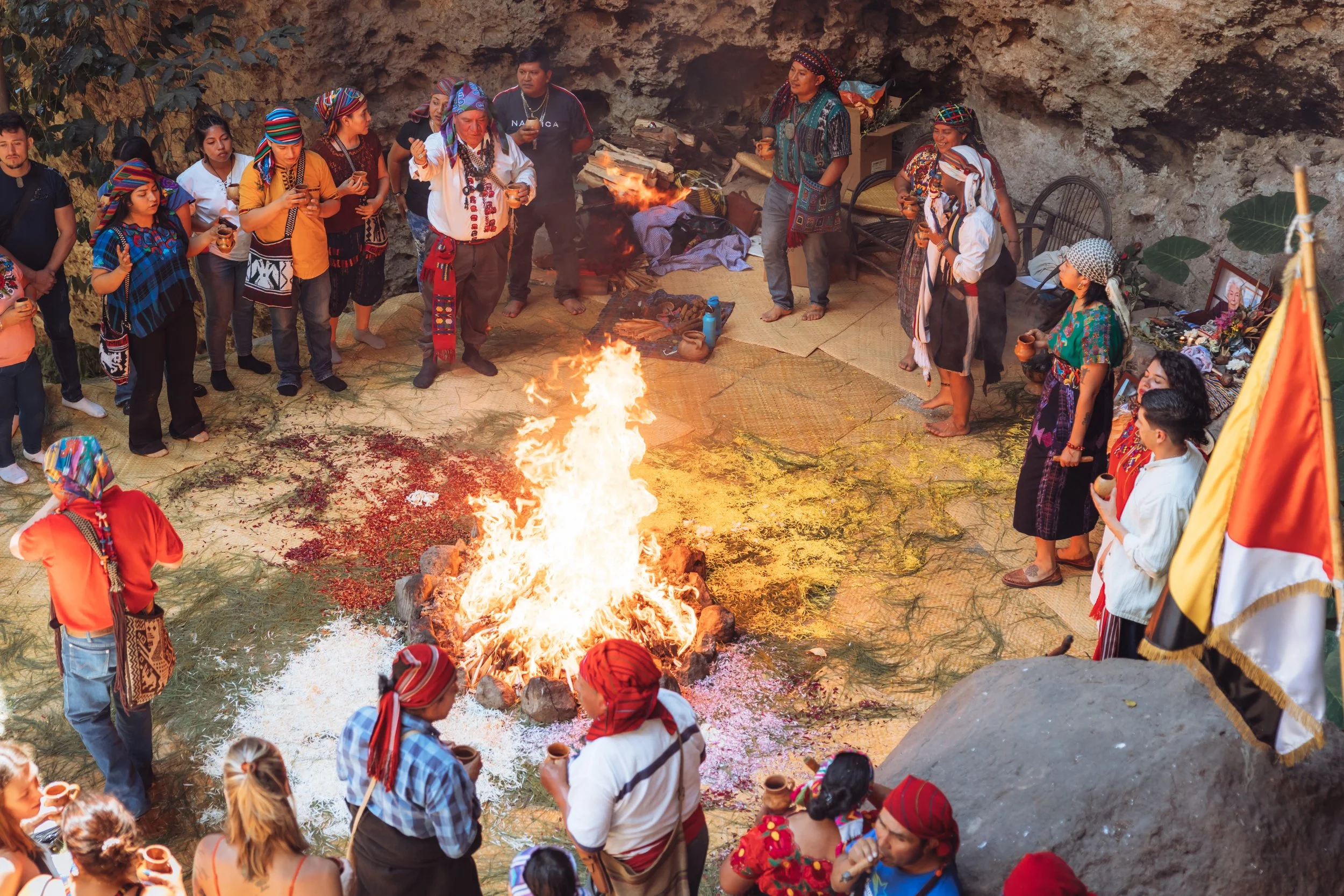 A group of people participating in a traditional ceremony around a large fire, many wearing colorful traditional clothing and headscarves, with some holding cups or bowls, in an outdoor setting with a rocky background.