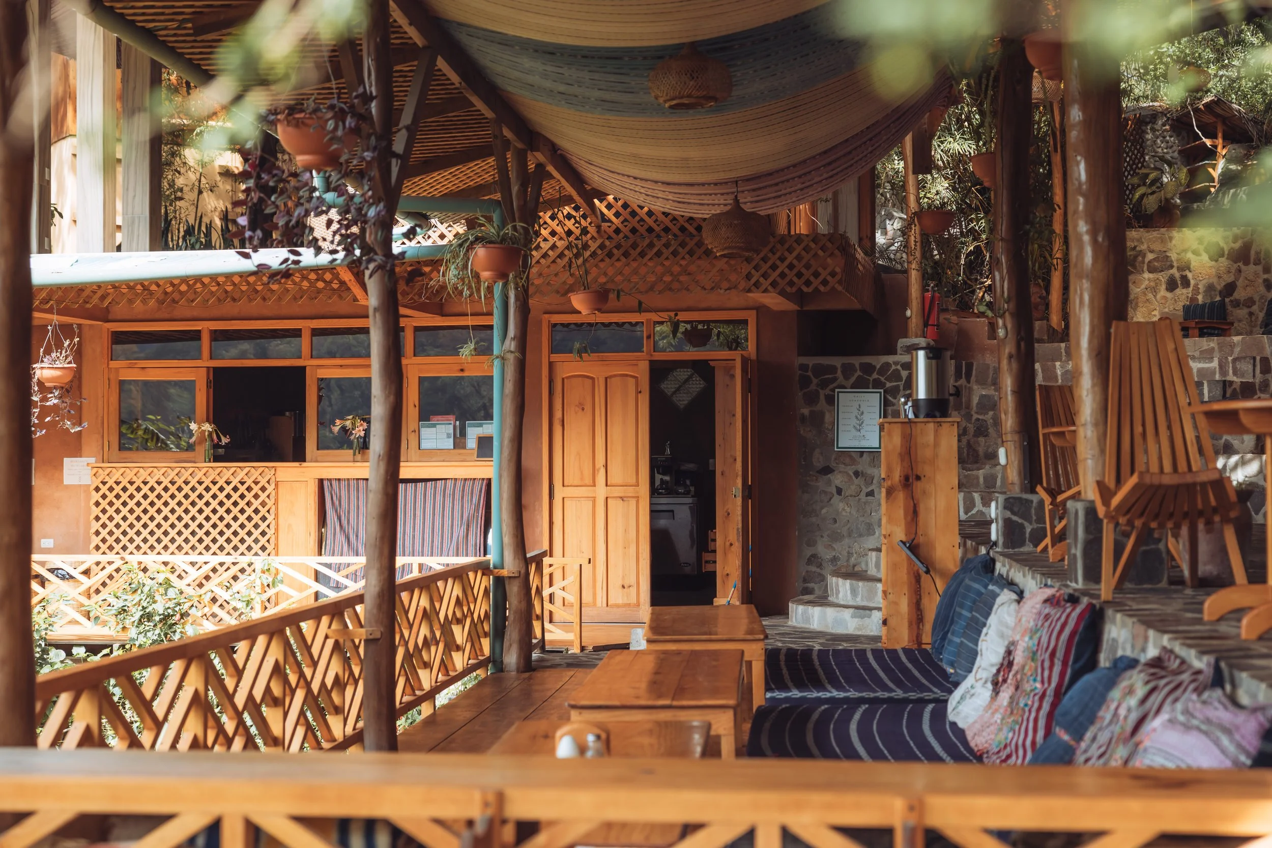 Wooden outdoor patio area with seating, hanging potted plants, and a stone wall in the background.