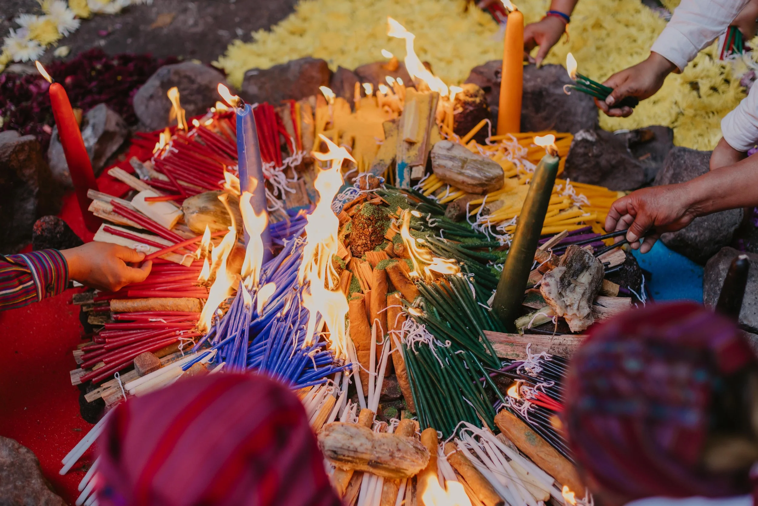 People lighting and arranging colorful candles with flames in a ceremonial setting, with rocks and yellow flowers in the background.
