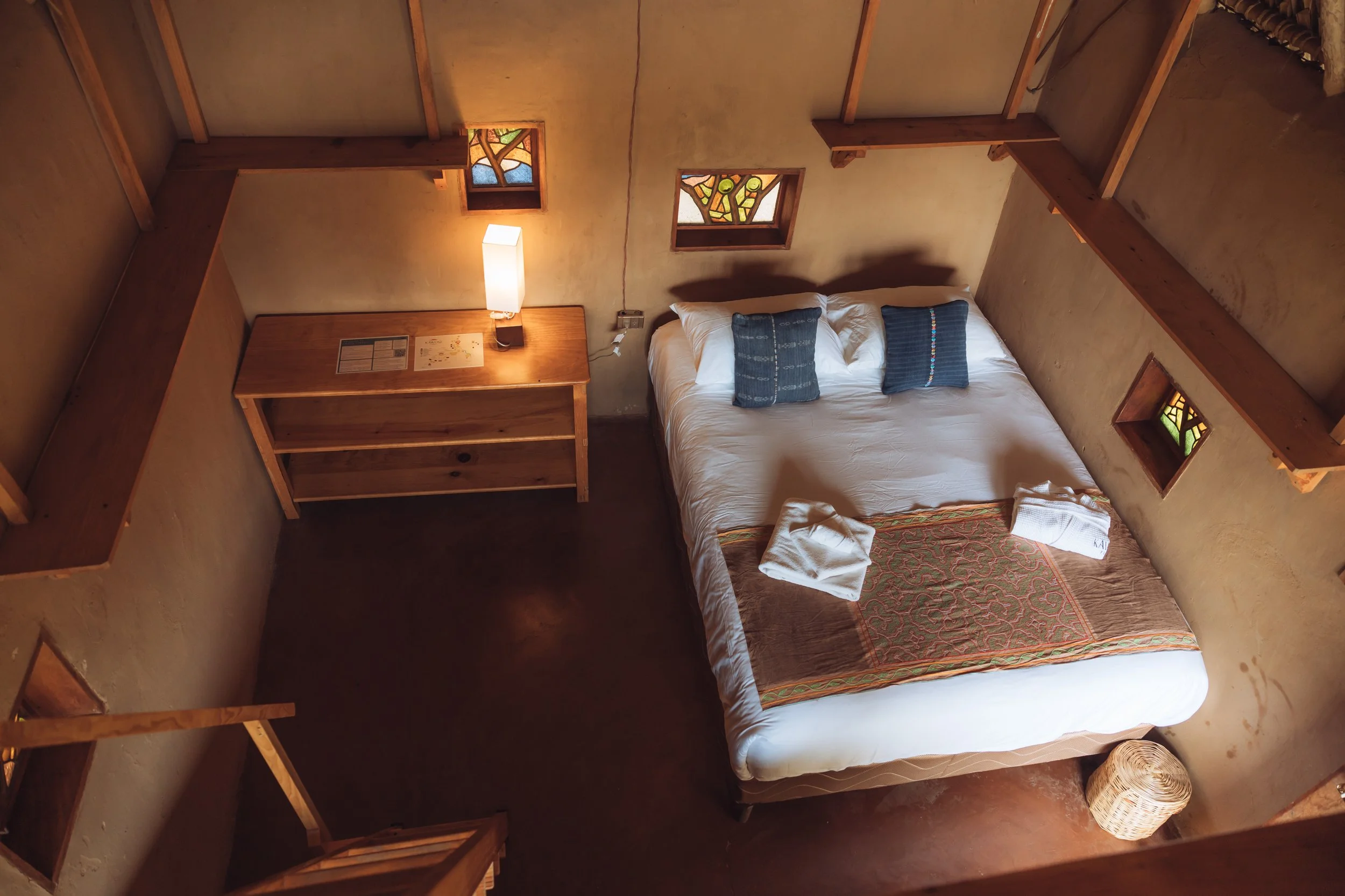 Top-down view of a cozy bedroom with a large bed, two pillows, two folded towels on the bed, small stained glass windows, a wooden dresser with a lamp, and a woven basket in the corner.