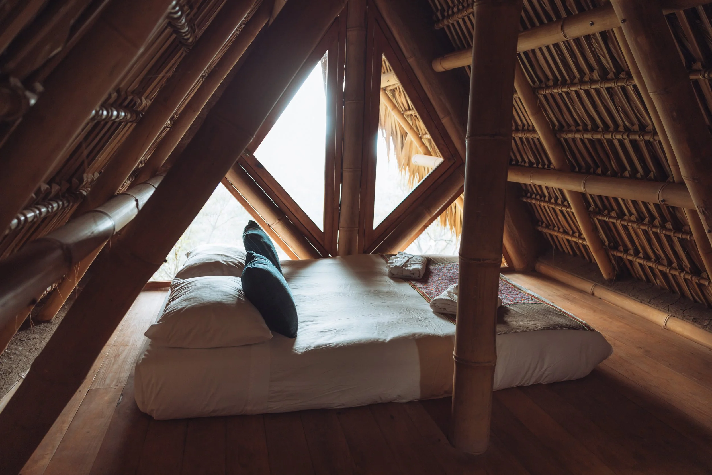 Interior of a rustic bamboo-straw hut with a low bed, white bedding, black pillows, and folded towels, with a large window showing outdoor scenery.
