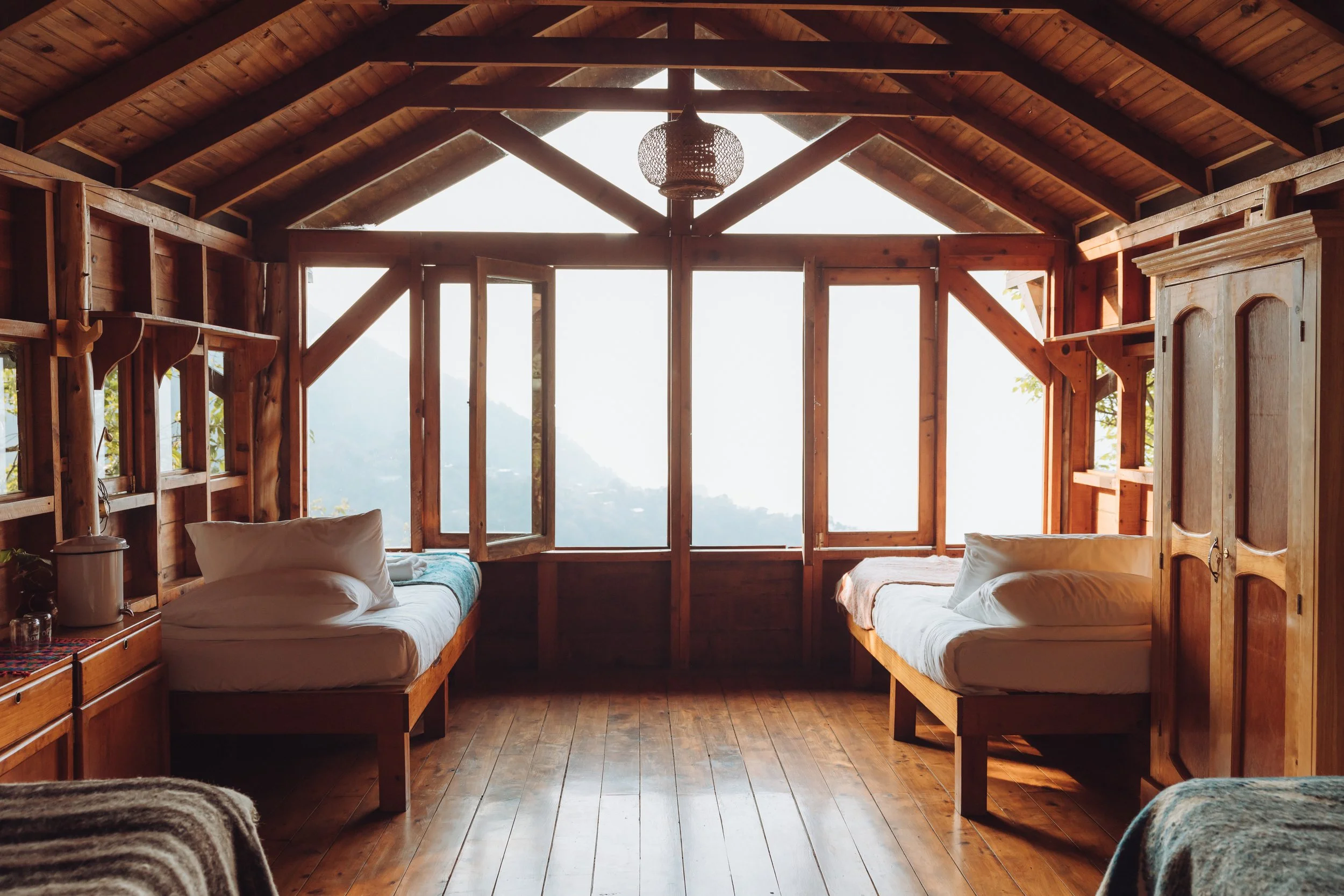 Two beds in a rustic wooden cabin with open windows, overlooking a mountain view.