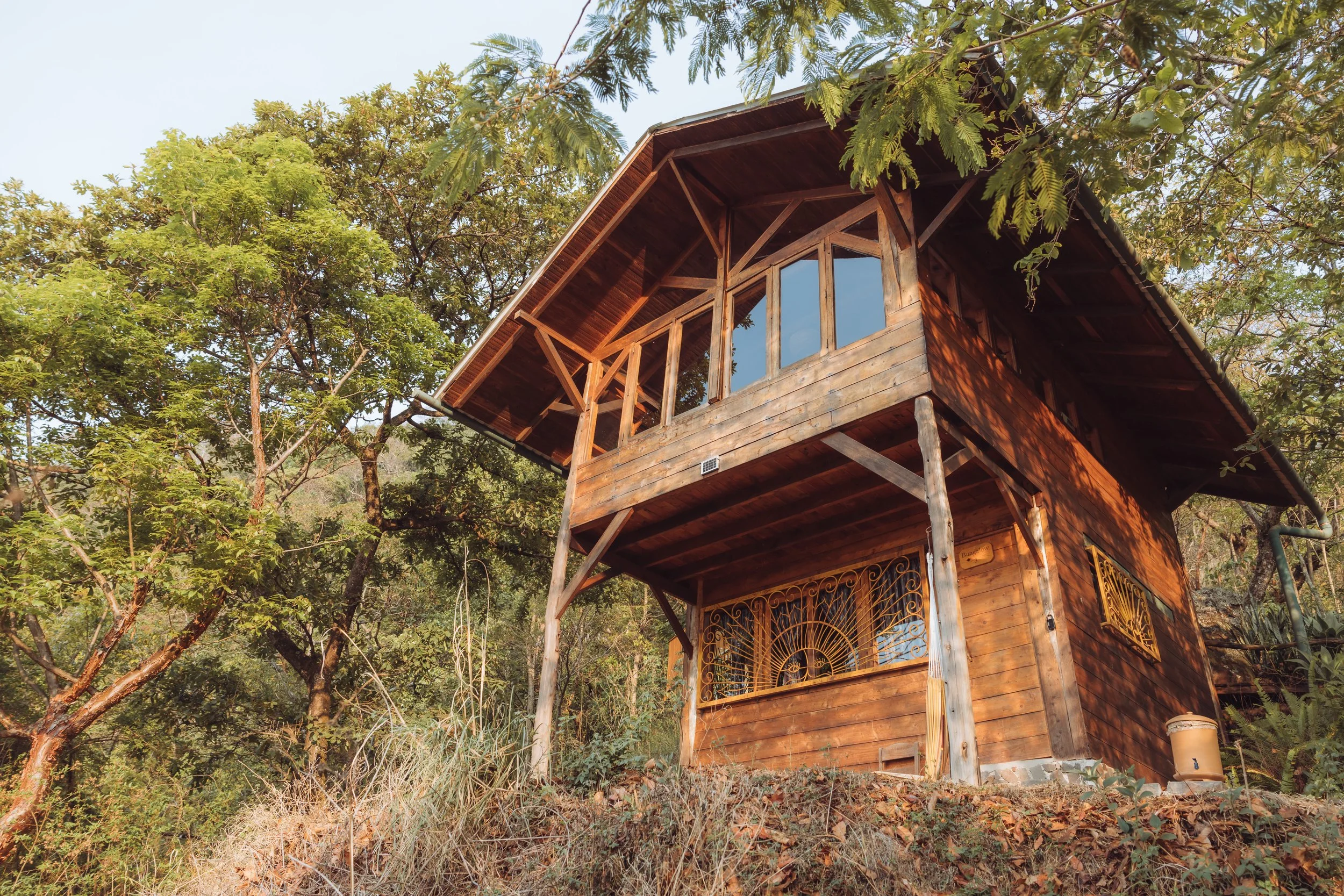 A wooden house on stilts surrounded by trees and greenery, with large windows and a balcony.