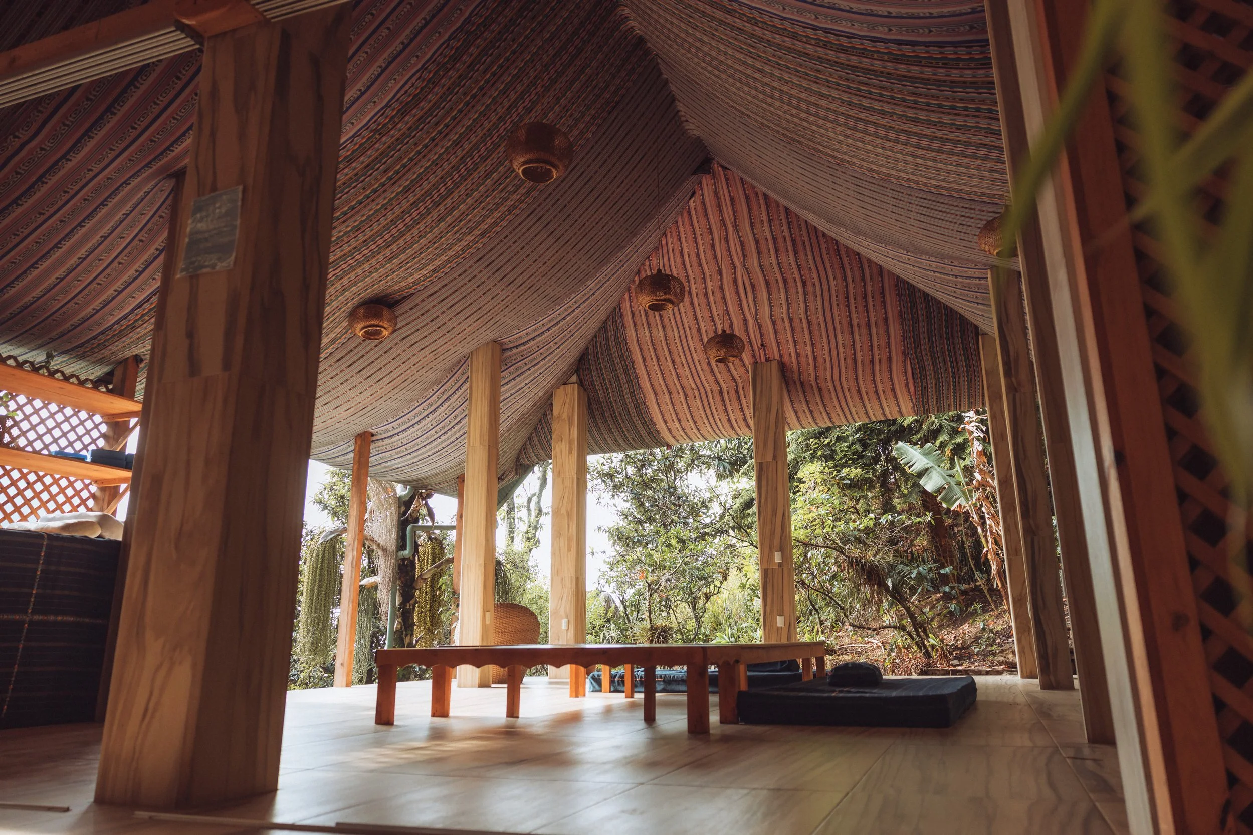 Open outdoor relaxation space with wooden pillars, patterned fabric ceiling, and view of trees and nature.