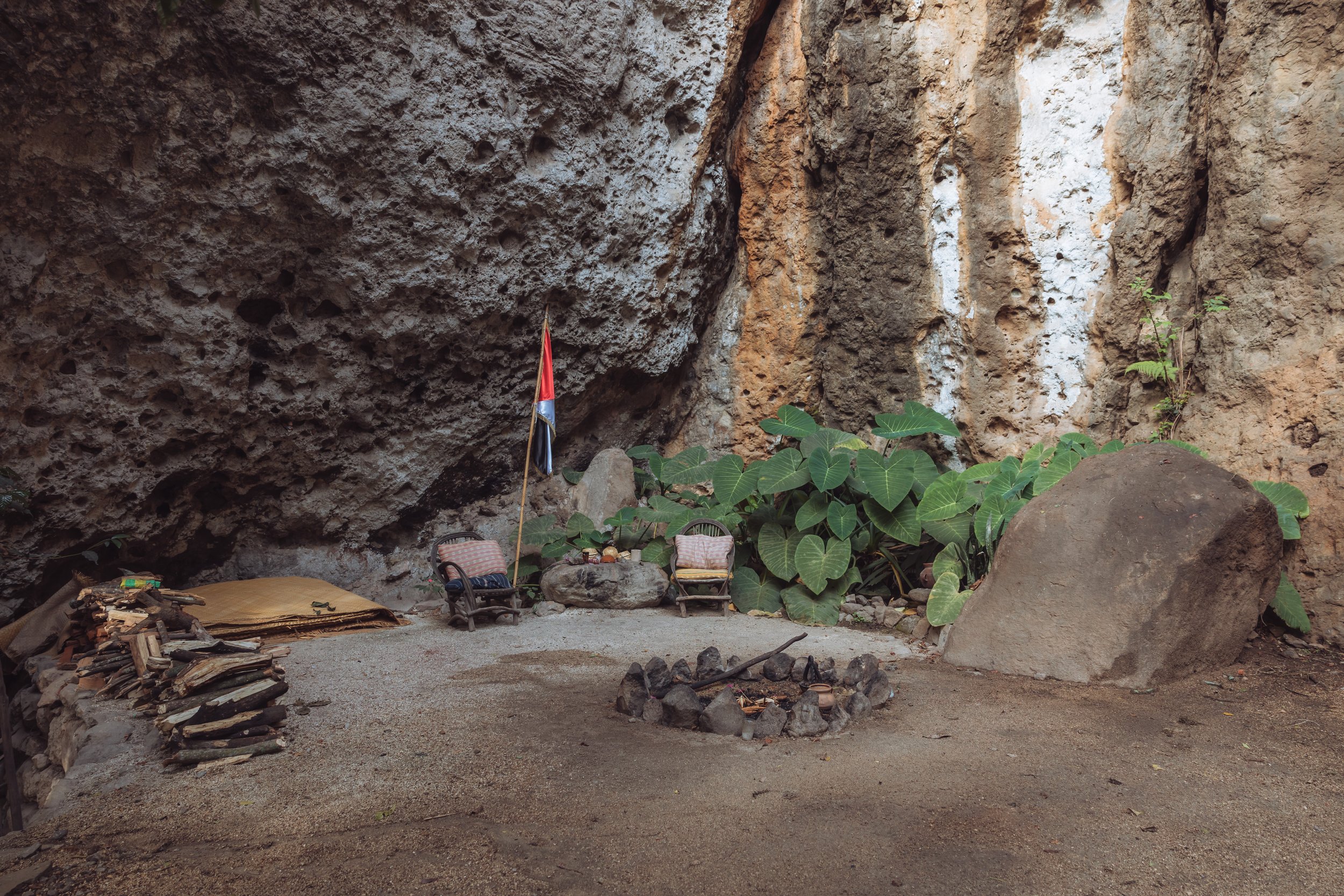 A small outdoor seating area inside a rocky cave with two wicker chairs, a flag, large green leafy plants, a large rock, and a circular stone fire pit.