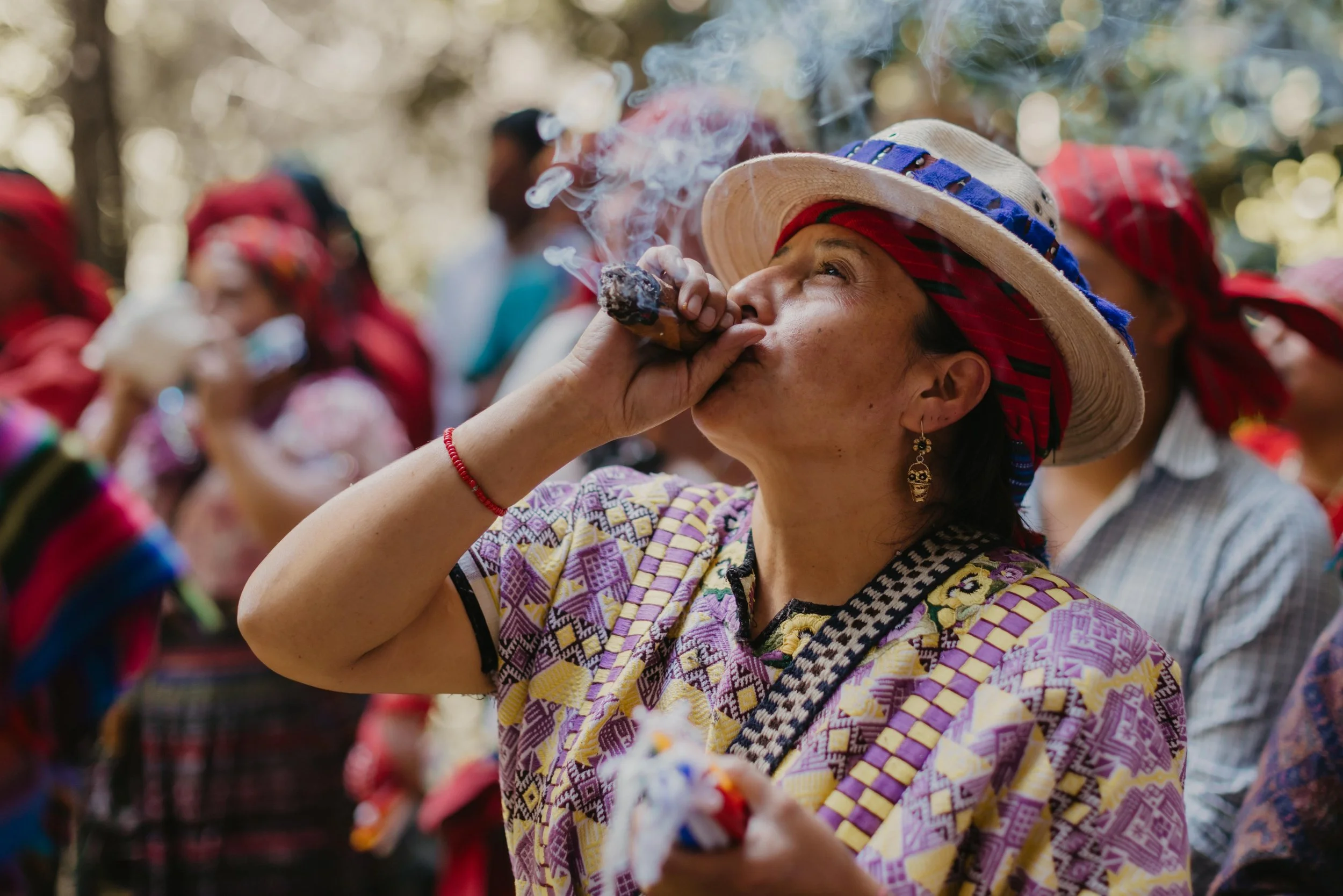 A woman from a traditional cultural gathering wearing a wide-brimmed hat and colorful clothing, smoking a cigarillo, with other women in similar attire in the background.