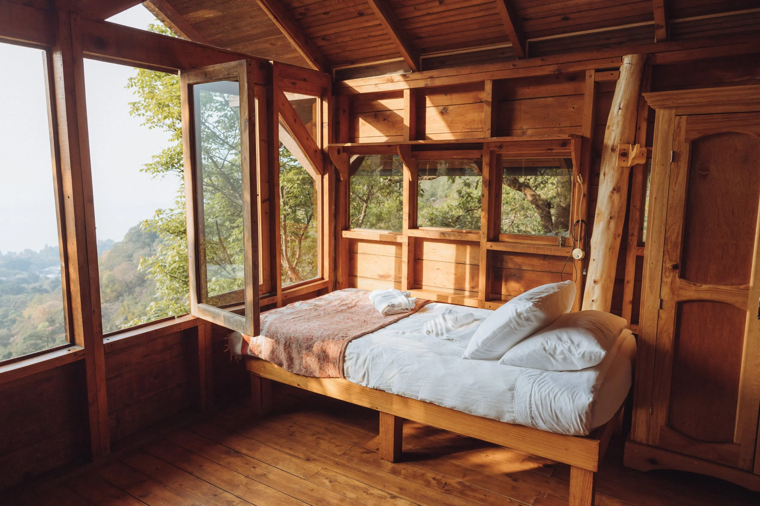 A cozy bedroom with wooden walls, large windows showing trees and nature outside, a bed with white linens and pillows, and sunlight streaming in.