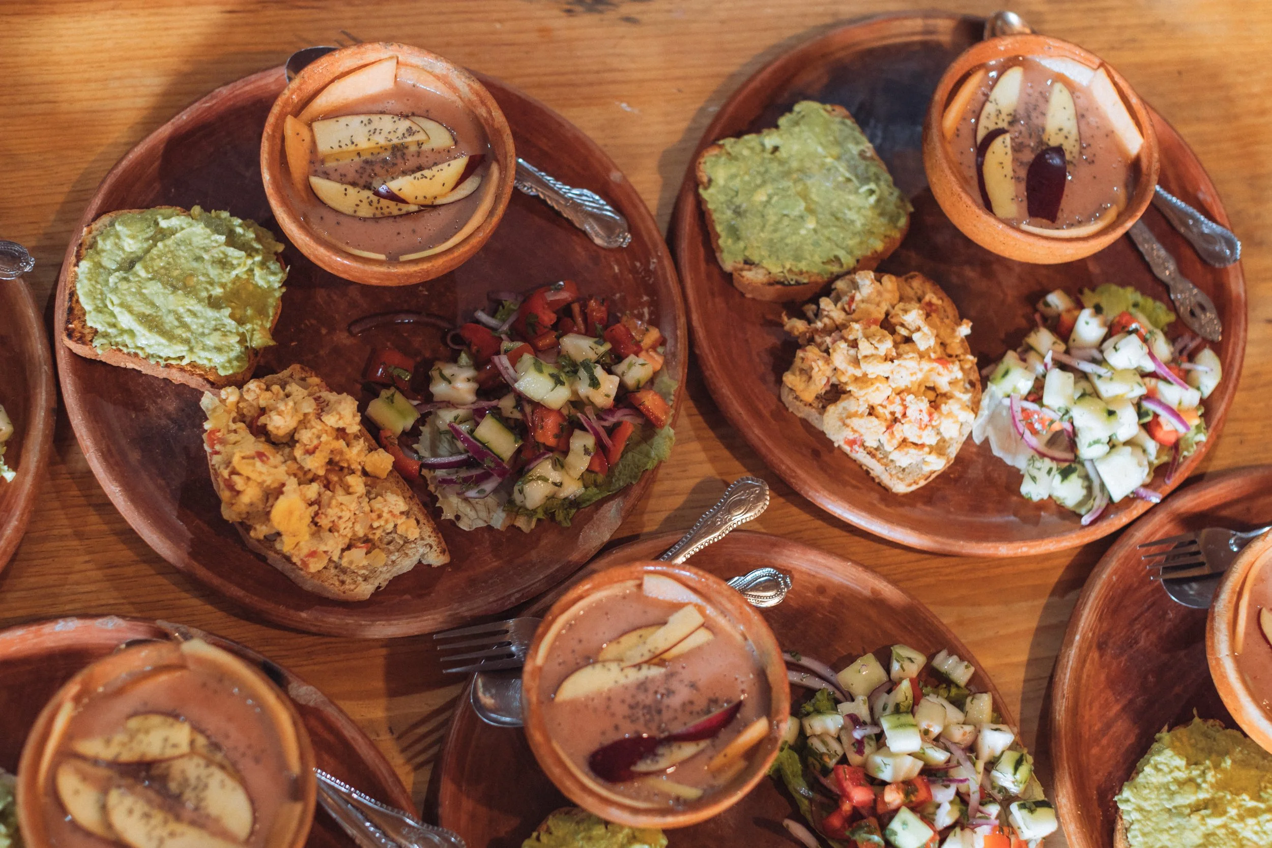 Multiple wooden plates with taco dishes, guacamole, and vegetable salads, accompanied by small bowls of fruit-based sauces or drinks, on a wooden table.