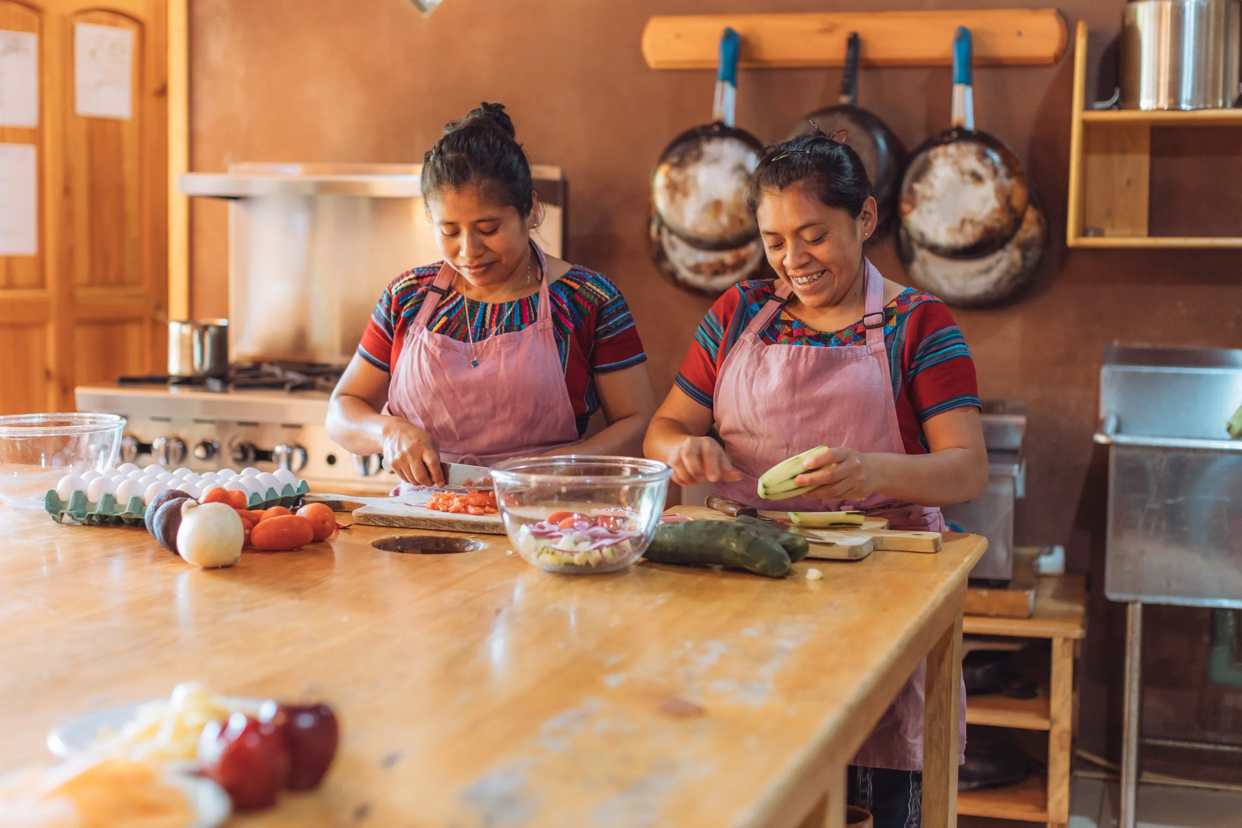 Two women wearing pink aprons and colorful traditional shirts chopping vegetables in a cozy kitchen with wooden shelves and hanging pots.