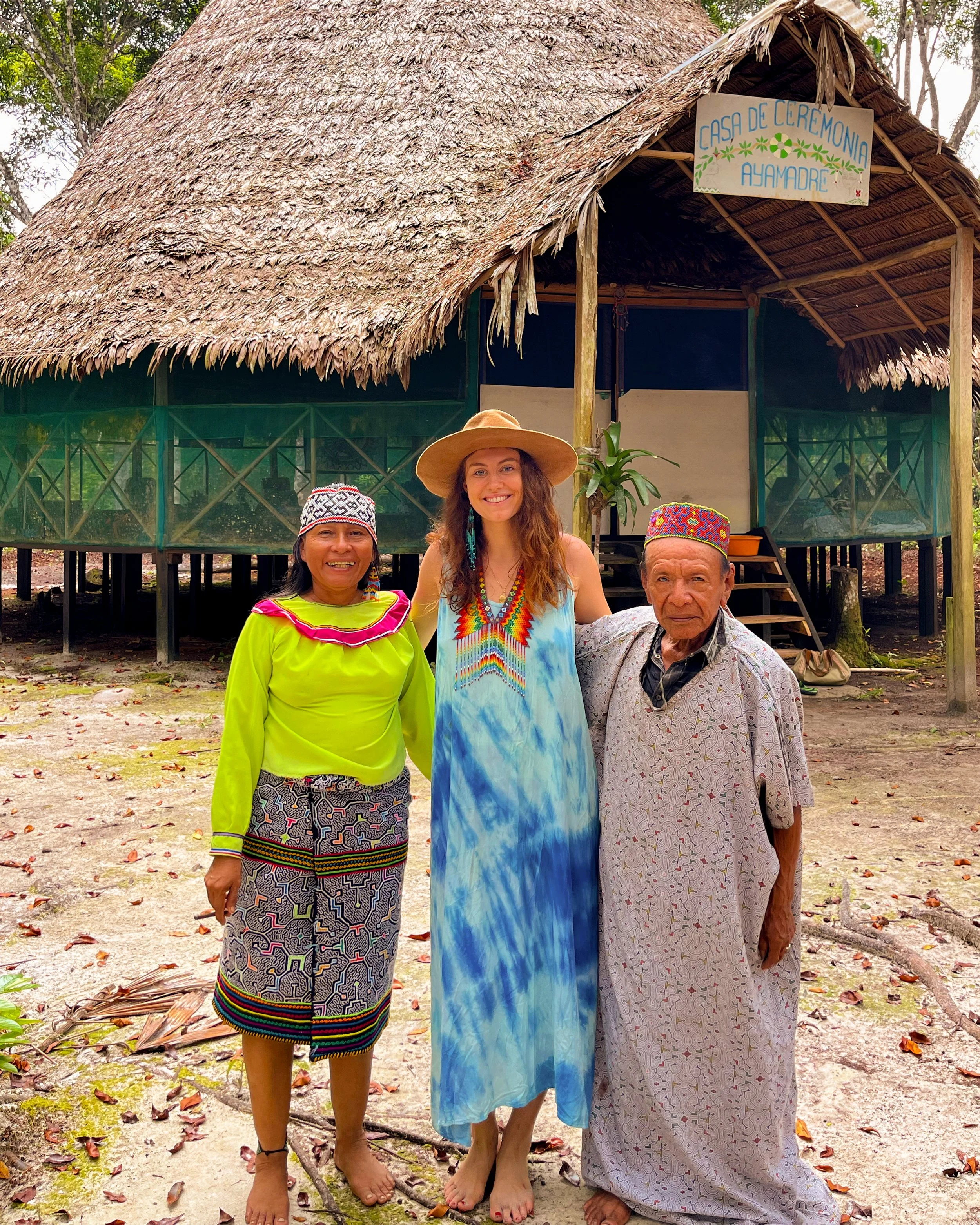 Three women standing together in front of a traditional thatched-roof building with a sign that reads 'Casa de Ceremonia Ayamadre' in a lush, green forest. The women are smiling and dressed in colorful, traditional clothing.