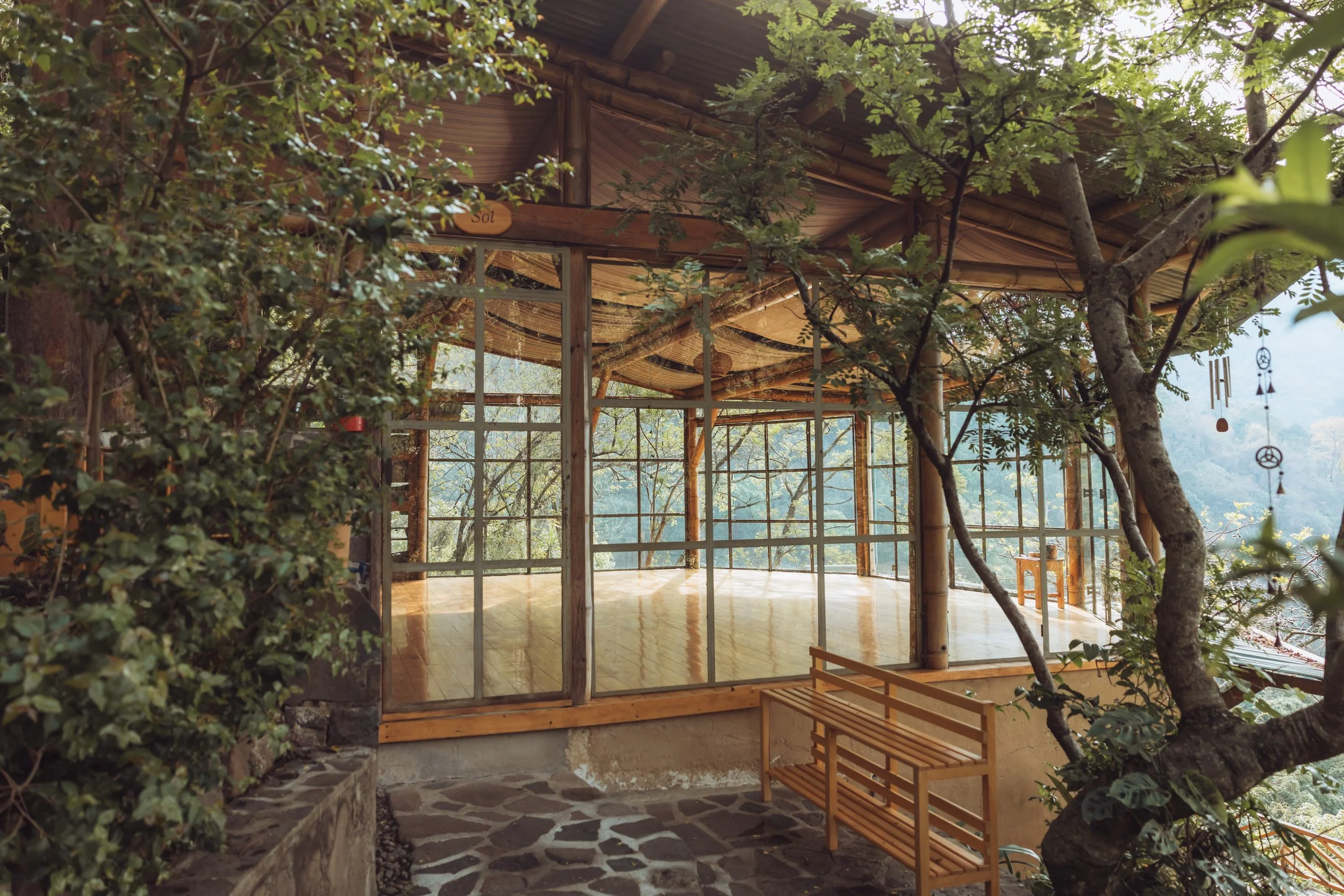 A rustic outdoor patio with a wooden floor, surrounded by trees and plants, enclosed with glass panels, overlooking a forested landscape, with wind chimes hanging on the right side.