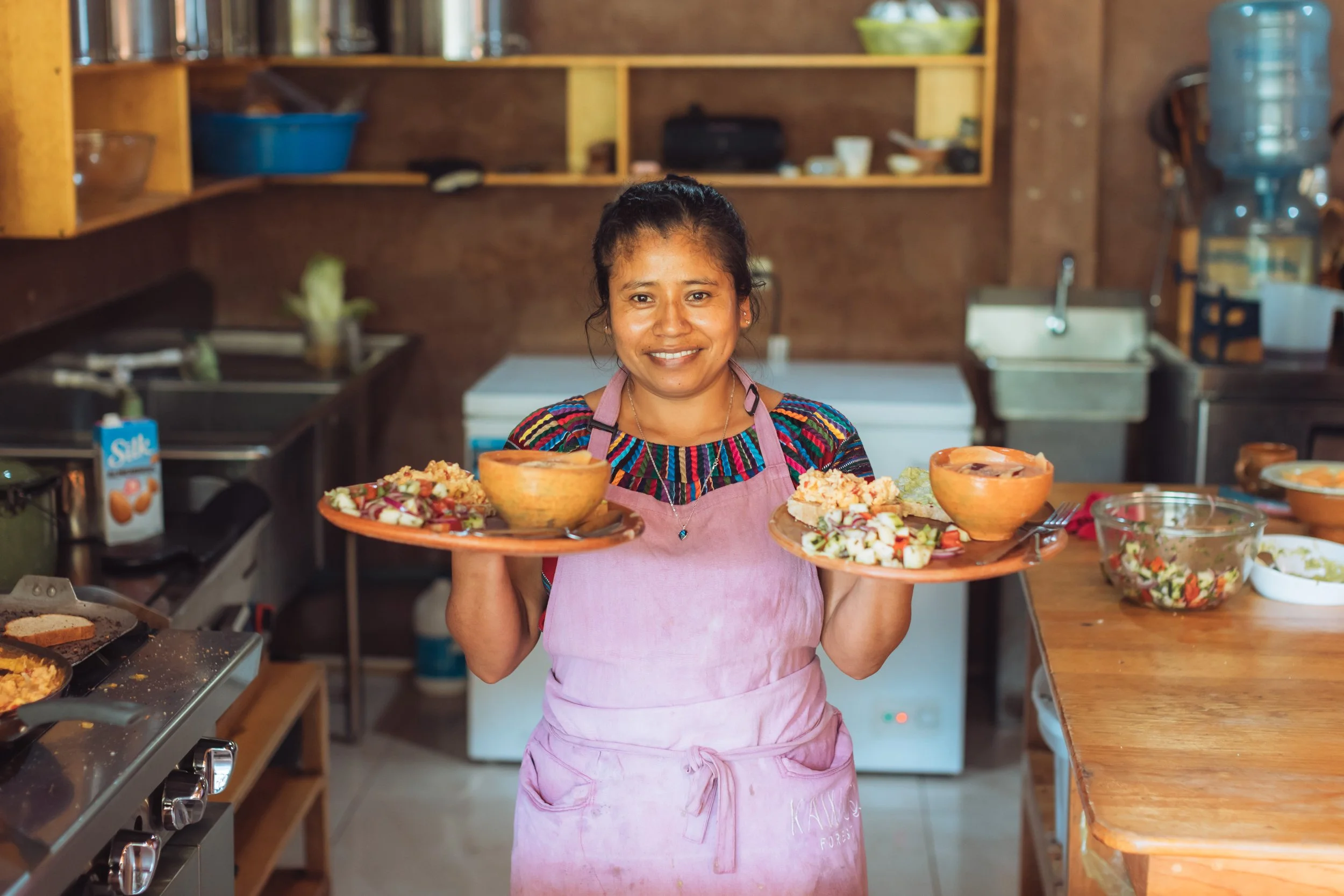 Smiling woman in apron holding two trays of food in a kitchen.
