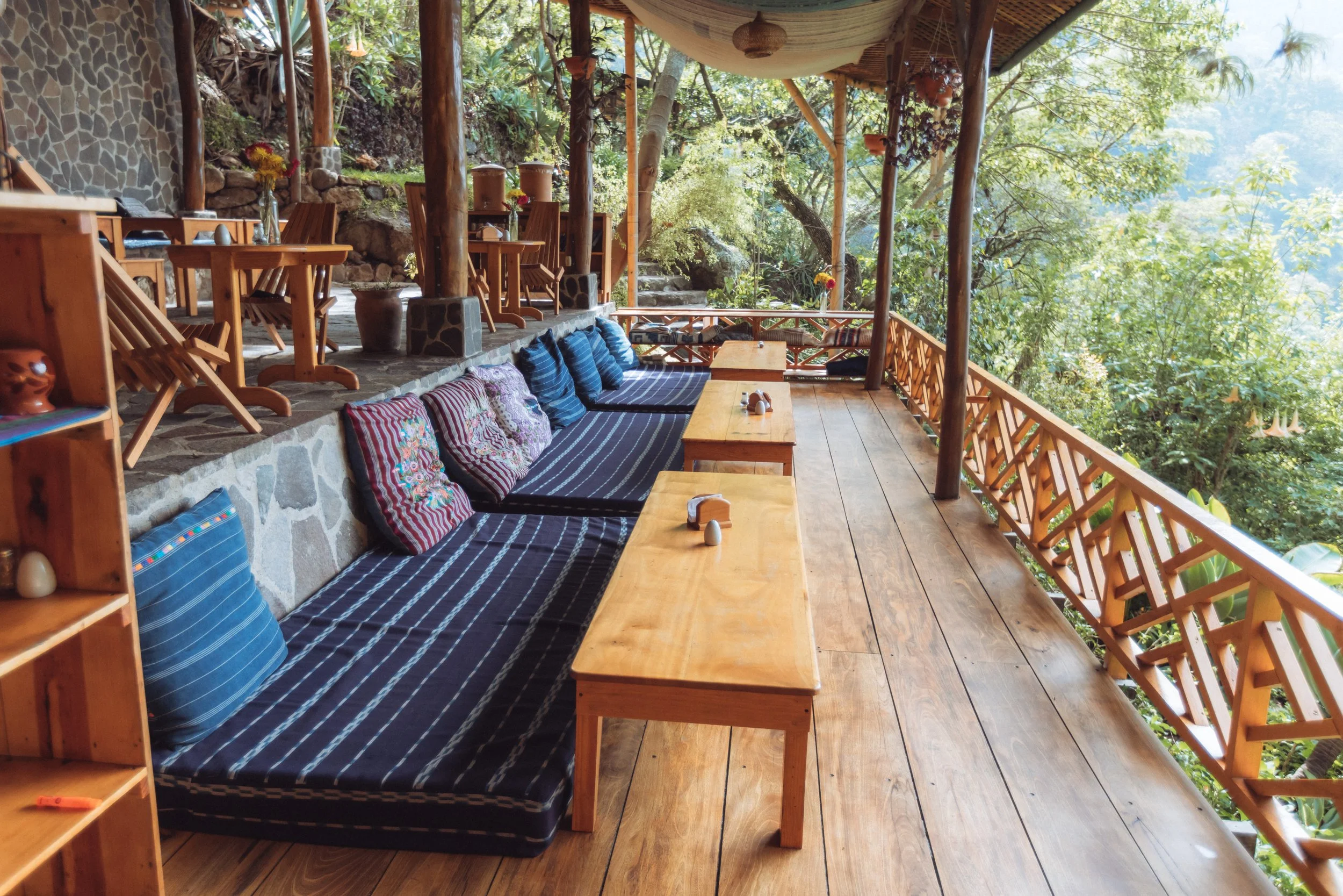 Wooden balcony with cushions, tables, and chairs surrounded by trees.