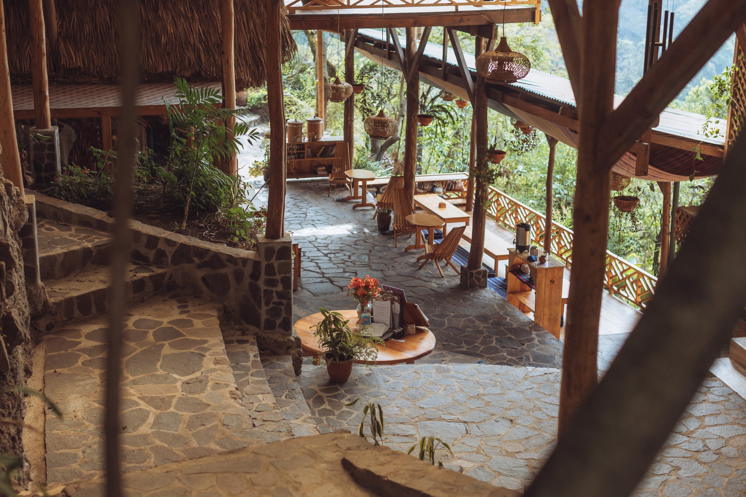 Interior of a rustic, open-air cafe or lounge with wooden furniture, stone flooring, hanging woven lamps, and surrounding greenery.
