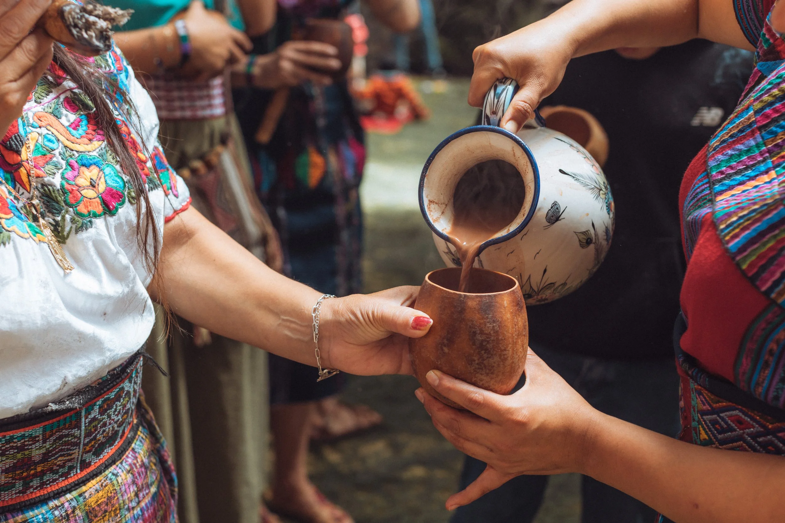 People in traditional Mexican clothing sharing and pouring hot chocolate from a decorated pitcher into a clay cup during a cultural event.