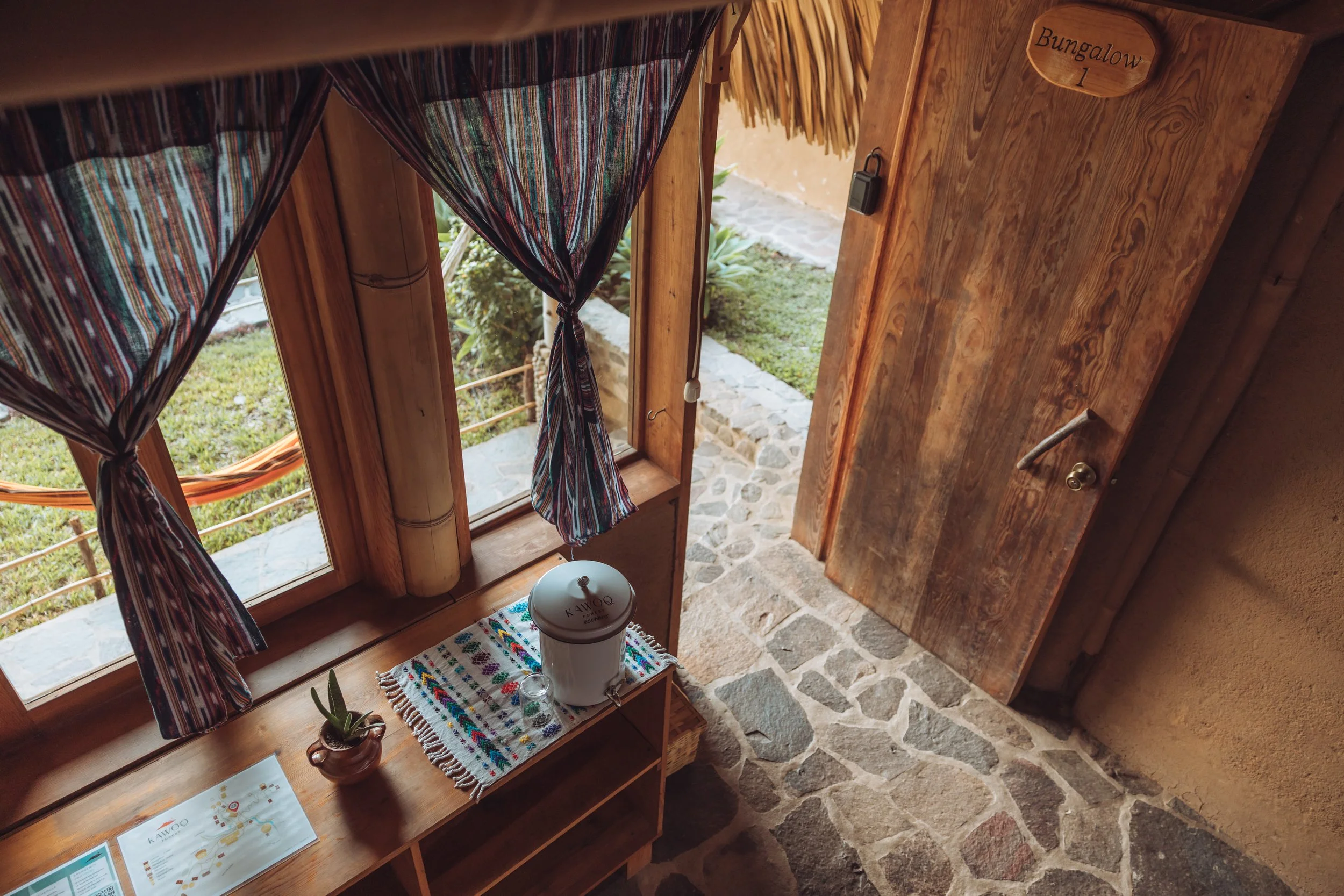 View of a rustic wooden door labeled "Bungalow 1," a small window with patterned curtains, a wooden shelf with a potted succulent plant and a white coffee dispenser, and a stone floor.