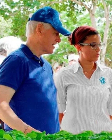 Older man and woman wearing glasses and casual clothing, standing outdoors amidst green trees, engaged in conversation.