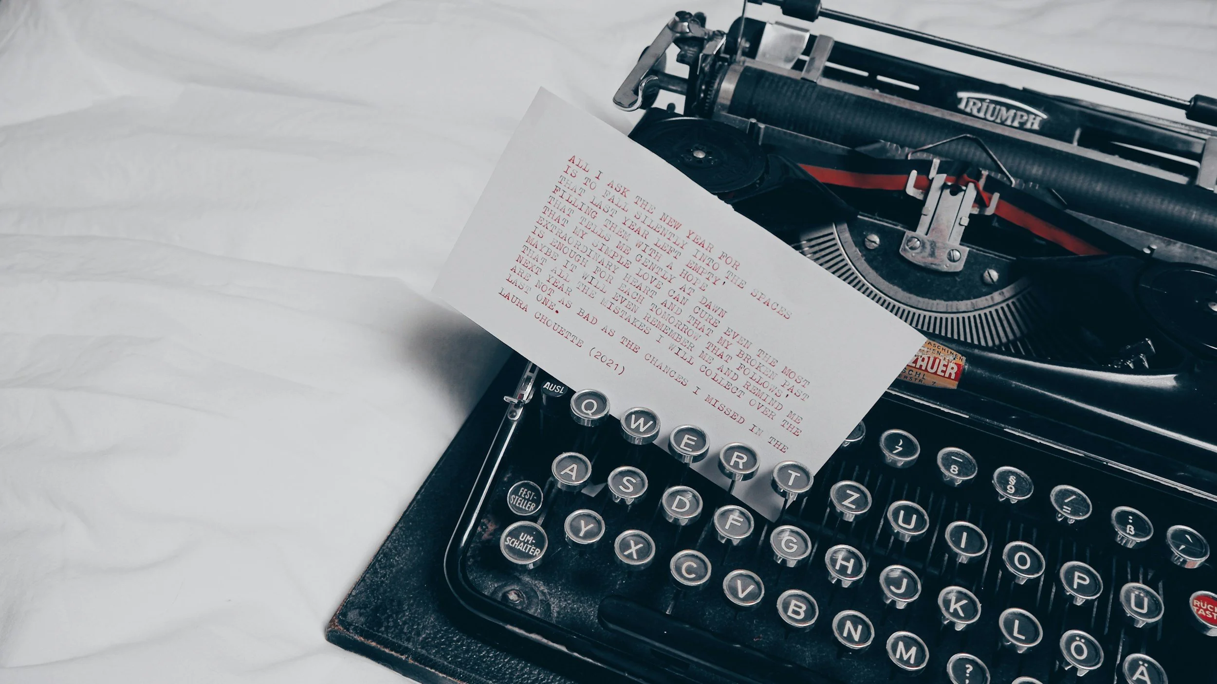 A close-up of a vintage black Triumph typewriter with a sheet of paper inserted, displaying typed red and black text.
