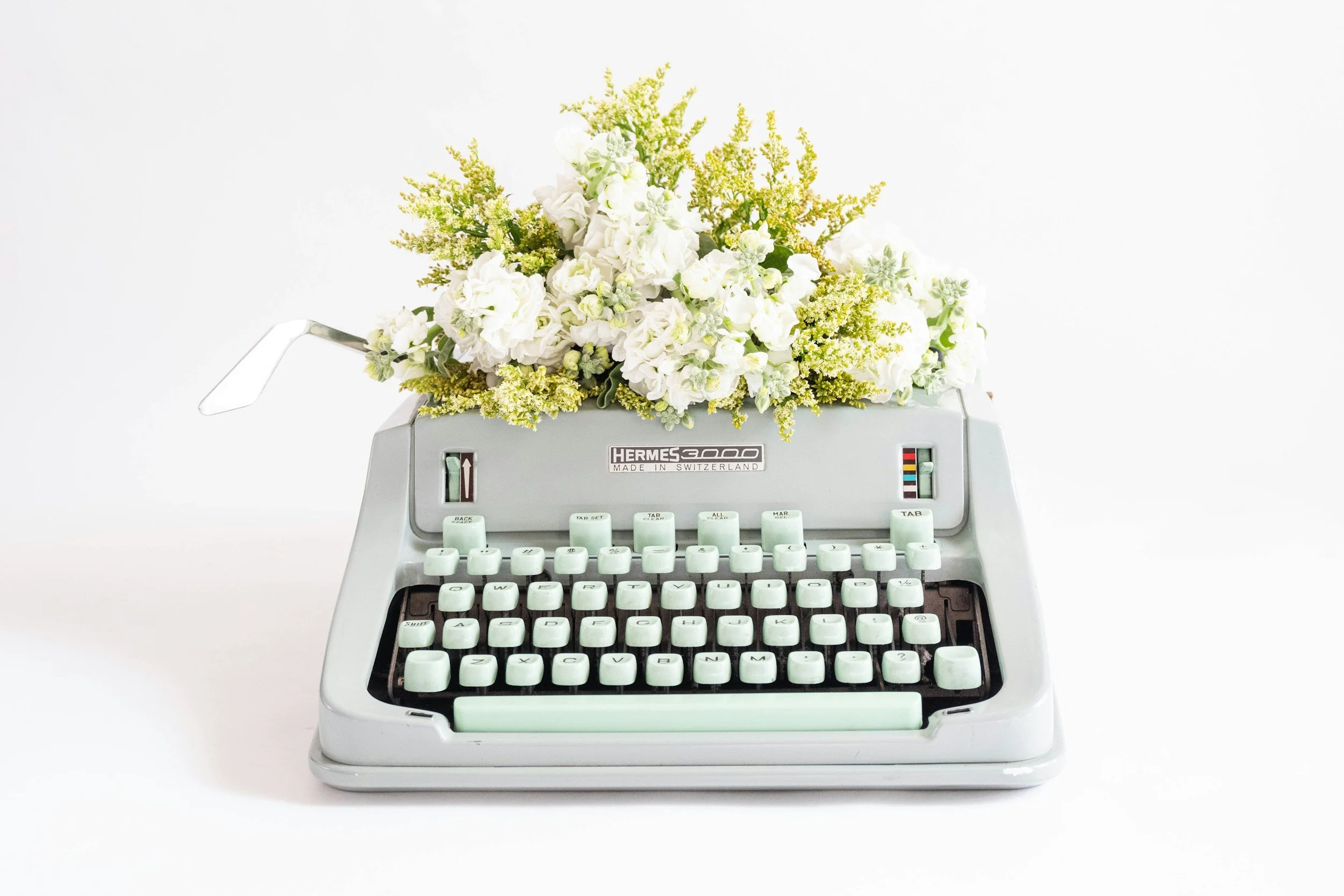 A vintage Hermes 3000 typewriter transformed into a flower arrangement with white and yellow flowers on a white background.