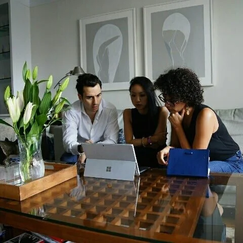 Three people seated around a wooden table looking at a Microsoft Surface tablet. Two women and one man, with a vase of white lilies on the table, in a modern interior with abstract wall art.