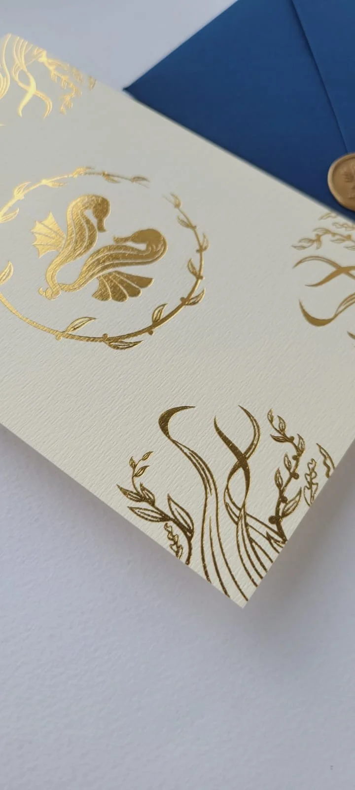 Close-up of a white card with gold foil designs featuring a seahorse and floral elements, accompanied by a partially visible blue envelope with a gold seal.