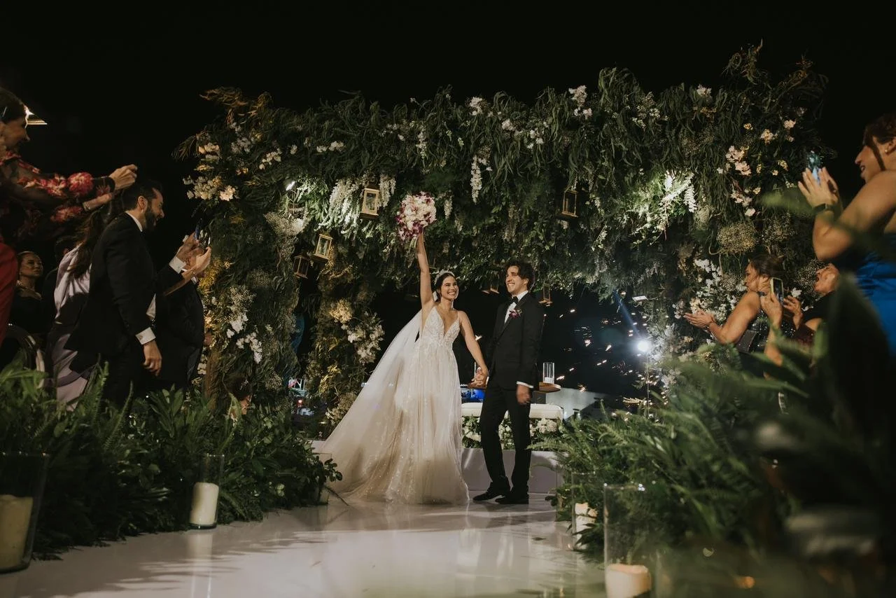 A bride and groom holding hands and smiling at each other under a floral arch at night during their wedding ceremony, surrounded by guests taking photos.