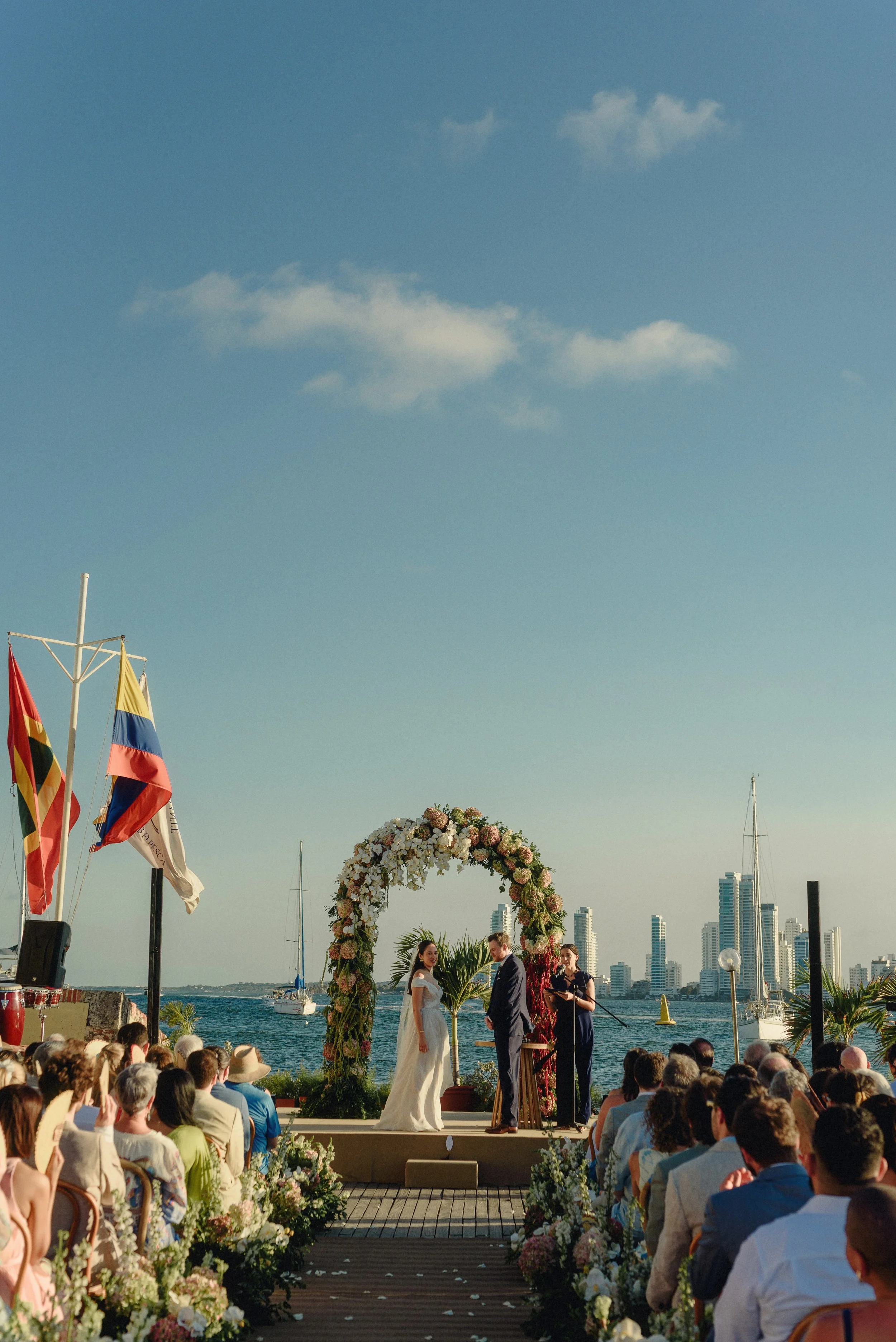 A wedding ceremony taking place outdoors by a waterfront with city skyline in the background. The bride and groom stand under a floral arch as officiant and photographer observe. Guests are seated facing the ceremony, and there are colorful flags and flower arrangements along the aisle.