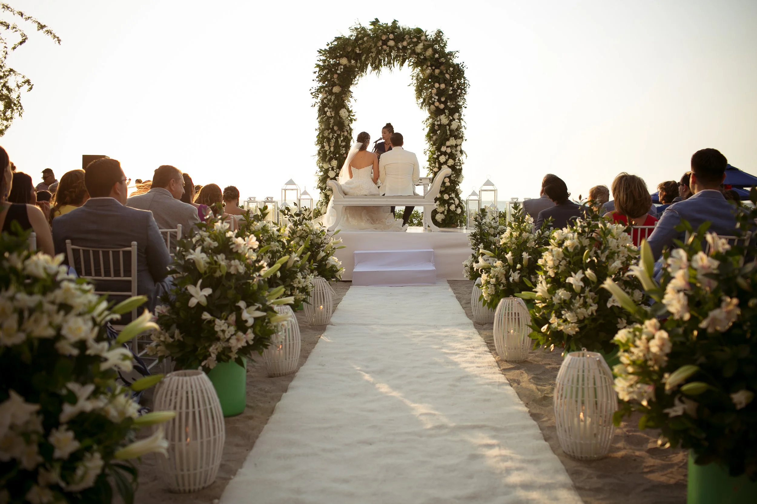 bilingual Wedding ceremony on a beach with a couple seated under a floral arch and guests seated on either side, decorated with white flowers and lanterns, during sunset.