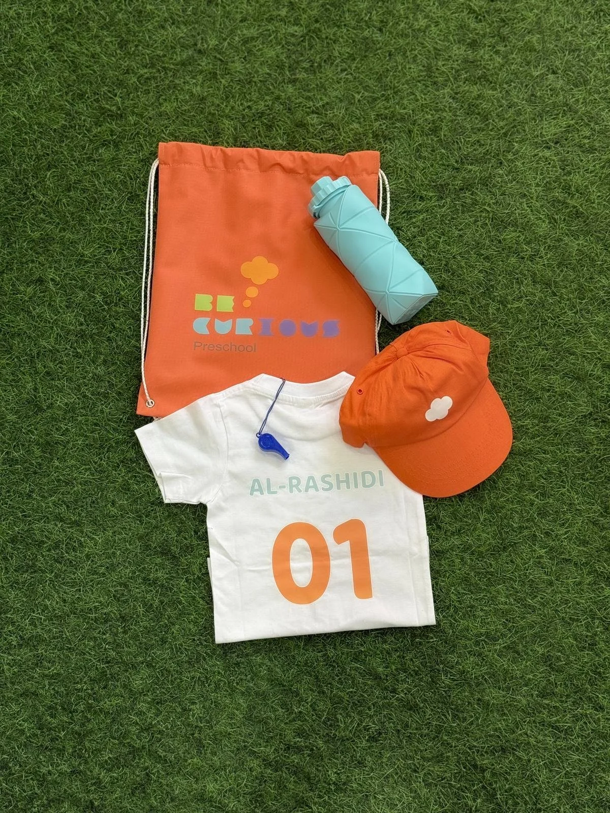 A white sports jersey with the name 'AL-RASHIDI' and number '01' in orange, a white whistle, an orange cap with a white cloud icon, an orange drawstring bag with 'BE CURIOUS Preschool' logo, and a mint green water bottle on green grass.