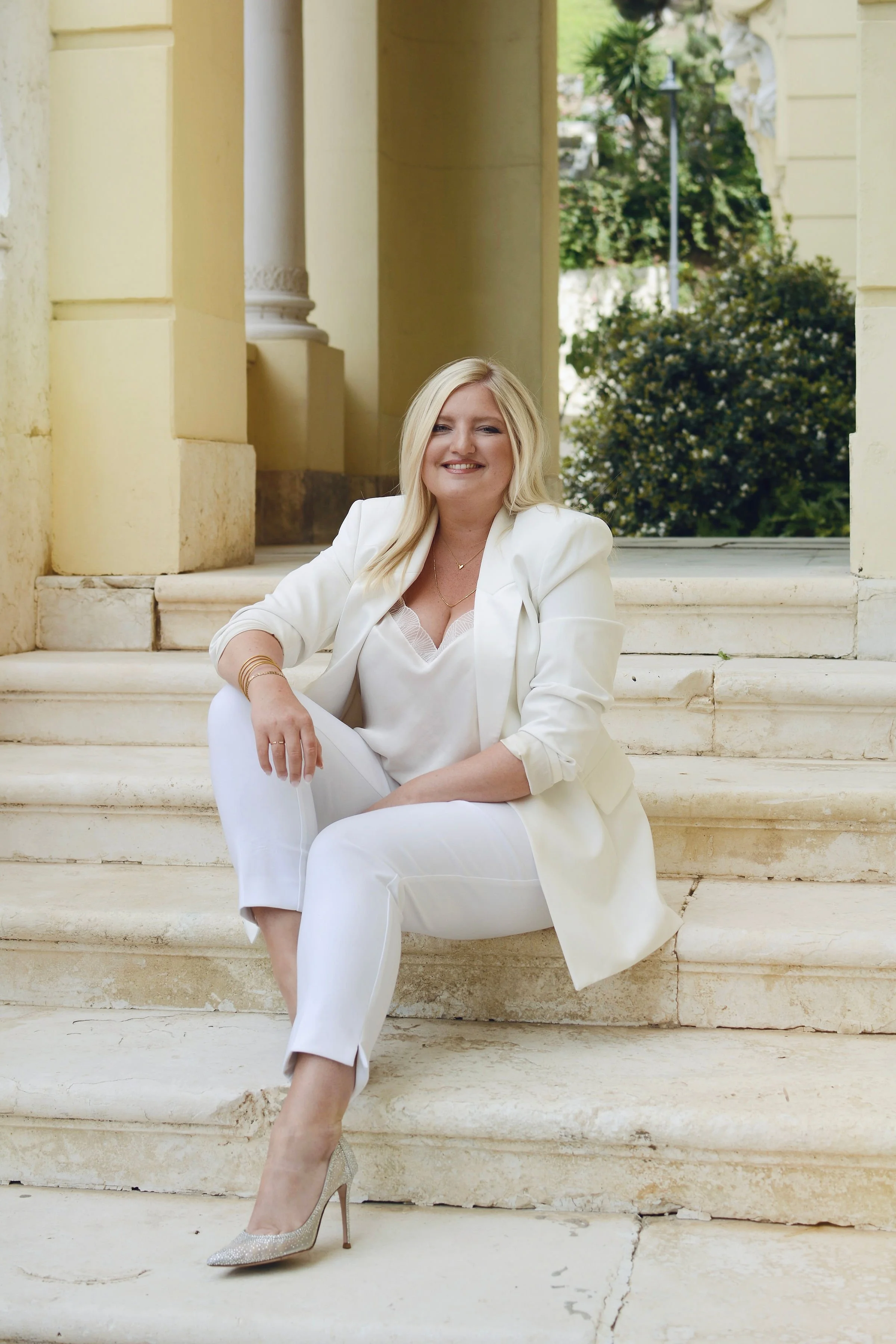 Maria, Founder of MVN Global in a white suit sitting on stone steps outdoors, smiling.
