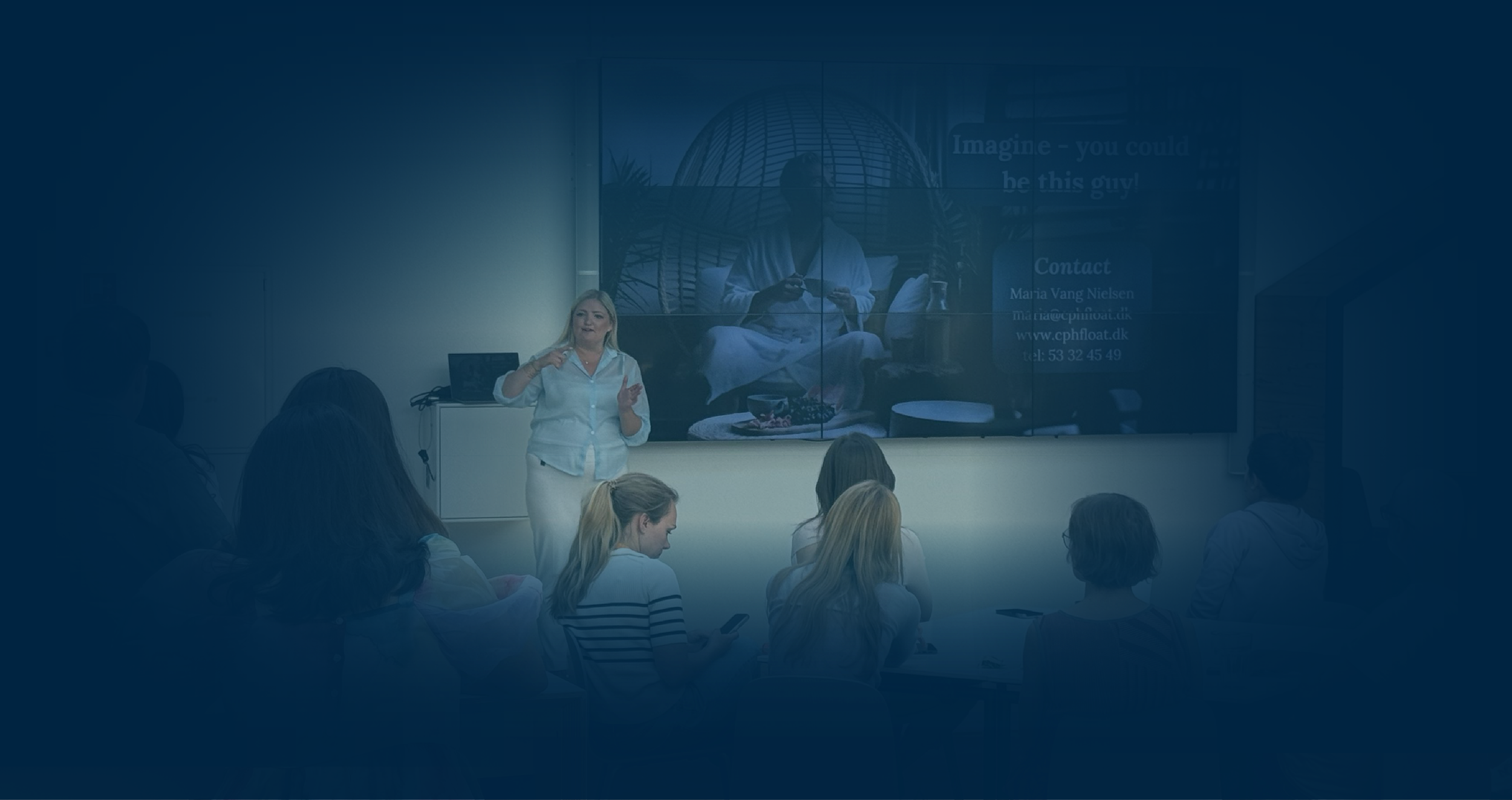 Maria, Founder of MVN Global is giving a presentation in a conference room, with an audience looking at her and a large screen displaying an image of a man sitting in a pod chair, holding a phone.