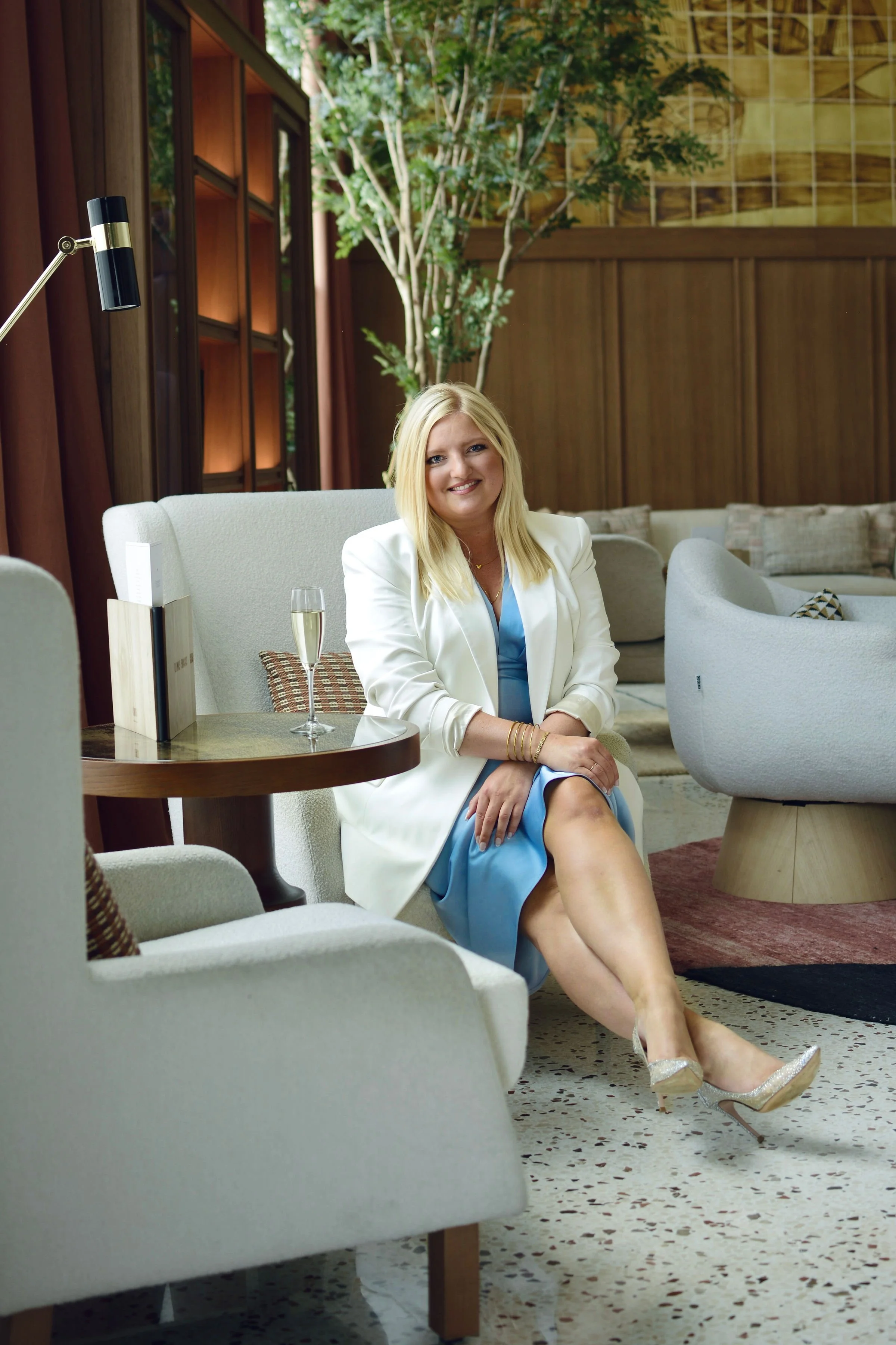 Maria, Founder of MVN Global woman with blonde hair wearing a white blazer and a light blue dress sitting on a beige armchair inside a well-lit lounge area with a glass of champagne on the side table.
