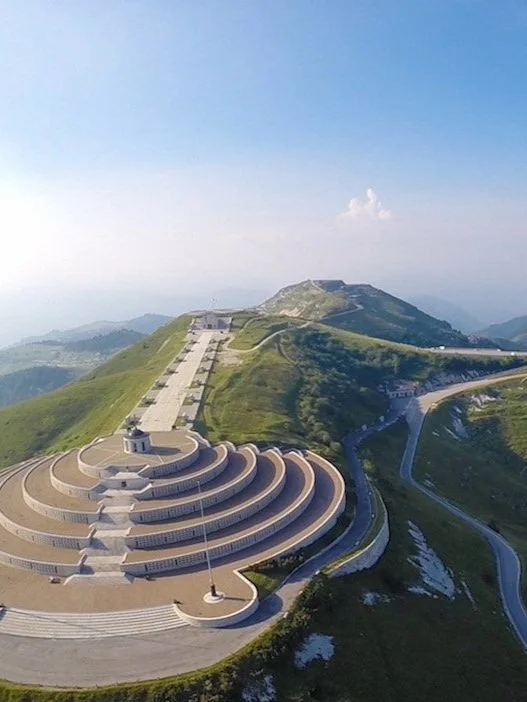 Panorama aereo del Sacrario Militare di Monte Grappa, con una struttura circolare a gradoni circondata da montagne verdi e un cielo azzurro.