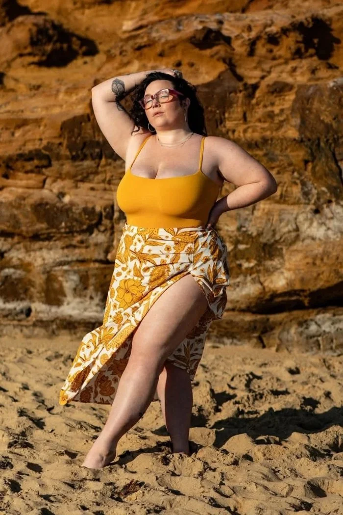 Woman with dark hair, wearing glasses, a yellow tank top, and a floral skirt with a high slit, standing on sandy ground against a rocky cliff background.