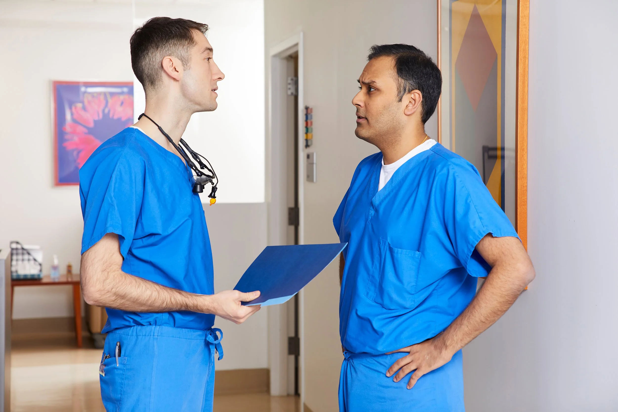 Two surgeons in blue scrubs talking in a bright hospital hallway with artwork on the walls. 