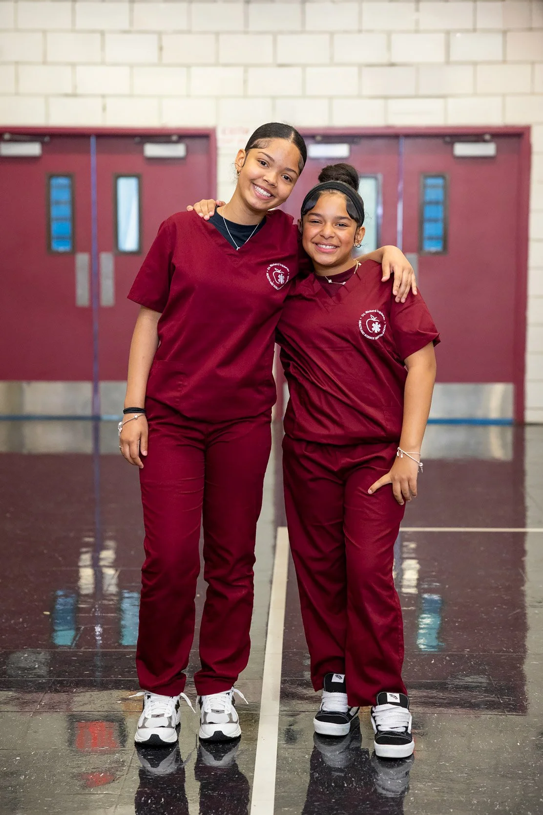 Two students standing with arms wrapped around each other in matching school uniforms in hallway.