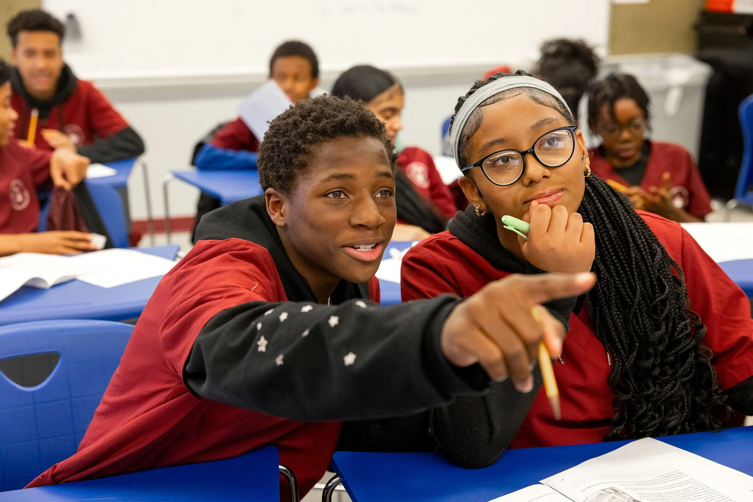 Two students in red uniforms smiling and pointing while working together in class.
