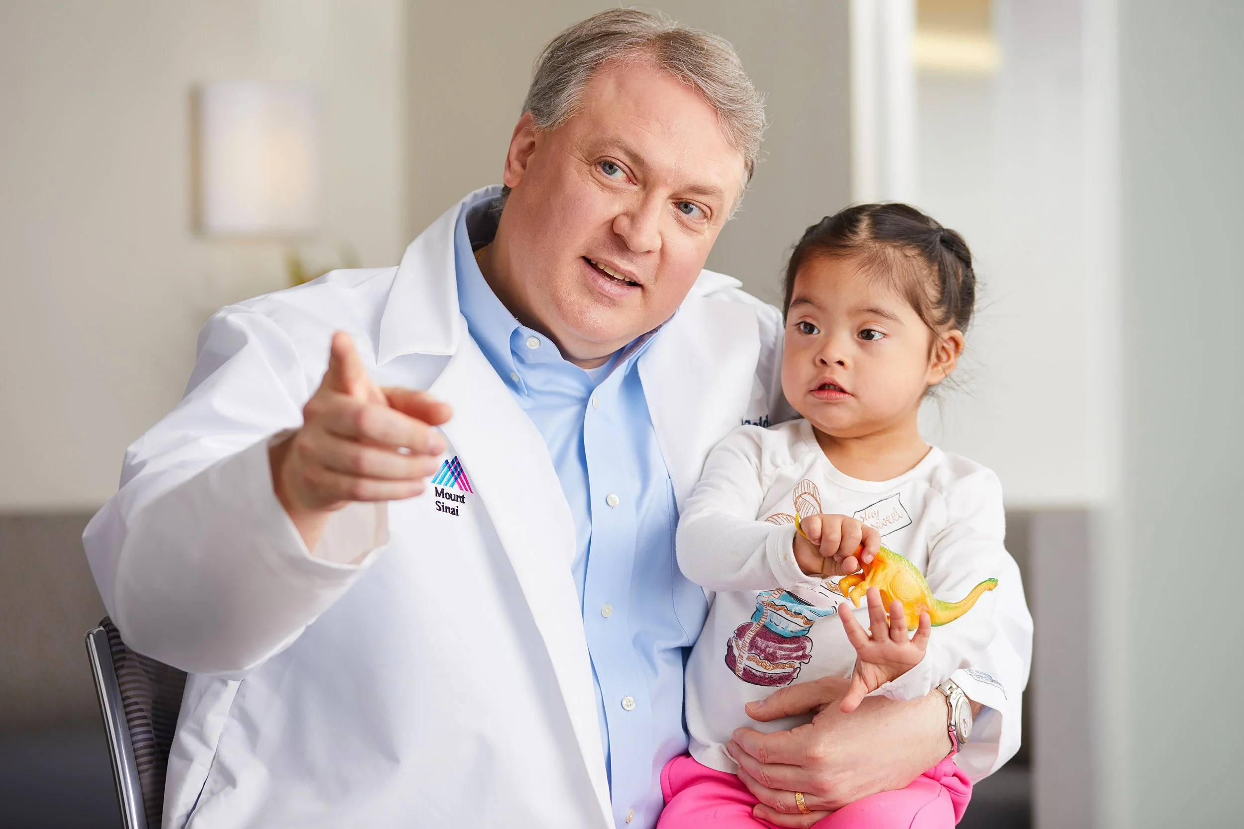 Doctor holding a young child patient with down syndrome while pointing towards the camera.