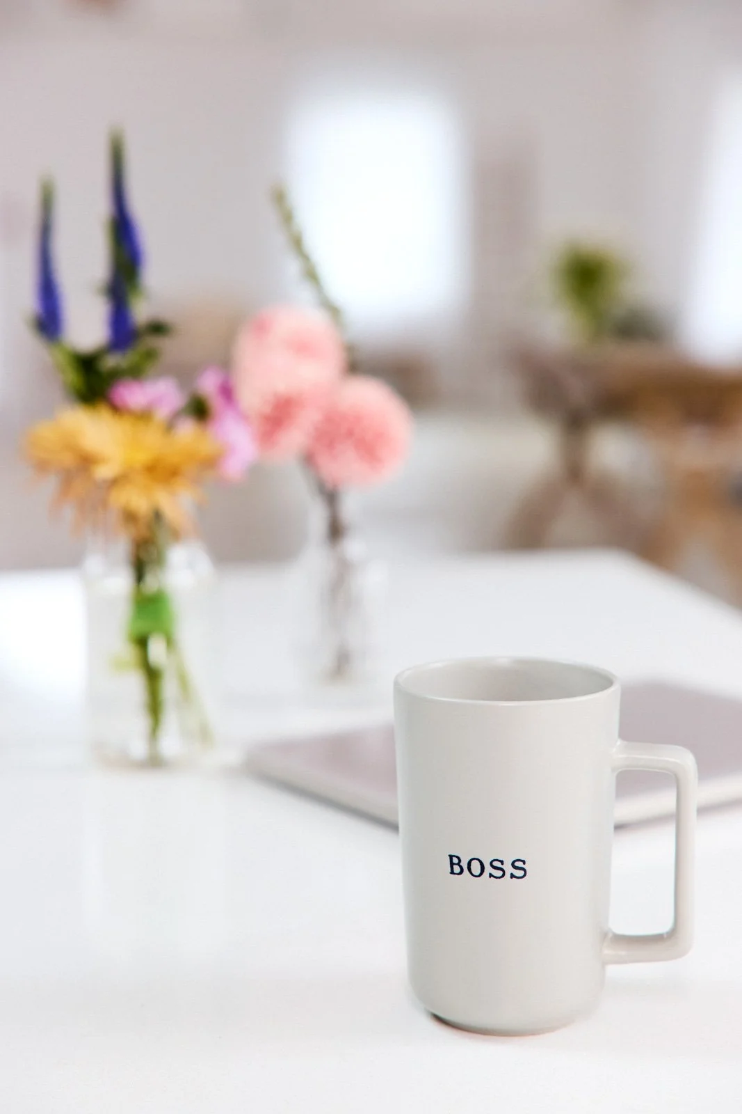 Neutral mug with BOSS inscription in front of a silver laptop on a white counter. Small vases with flowers in the background in a wide open space with shallow depth of field.