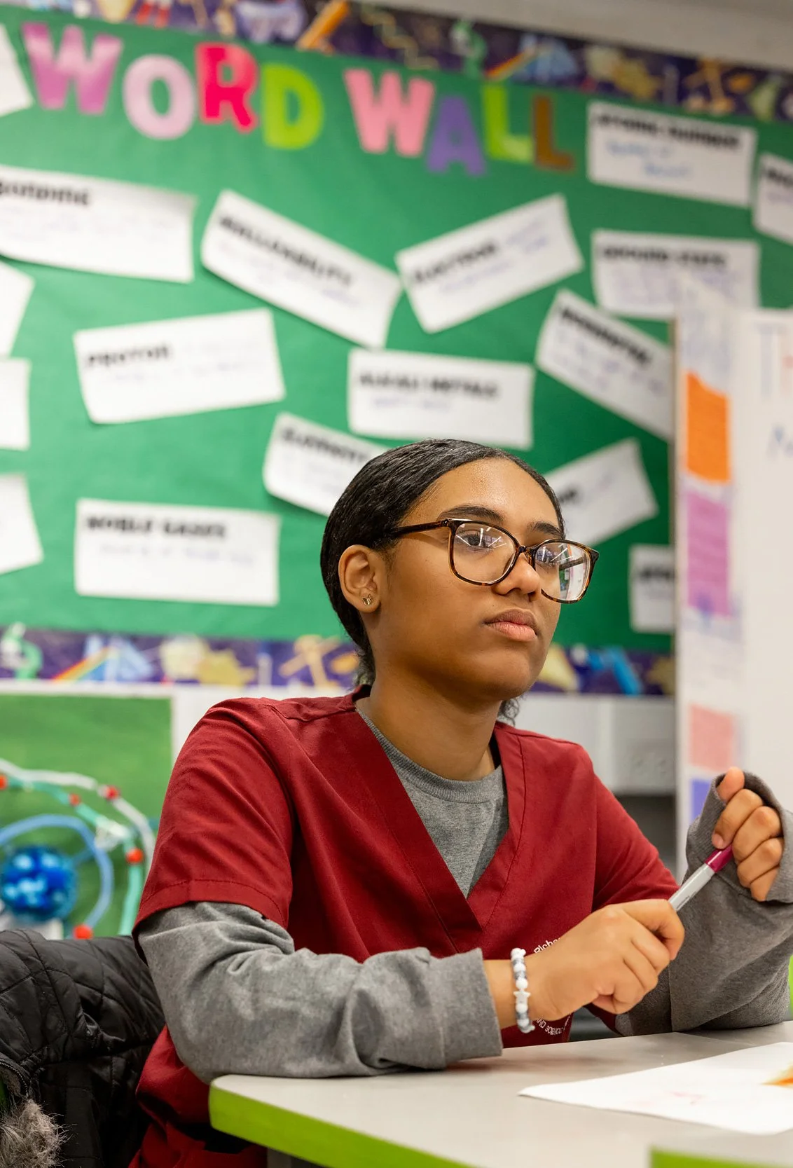 Student with glasses sitting in classroom with educational posters on wall behind them.