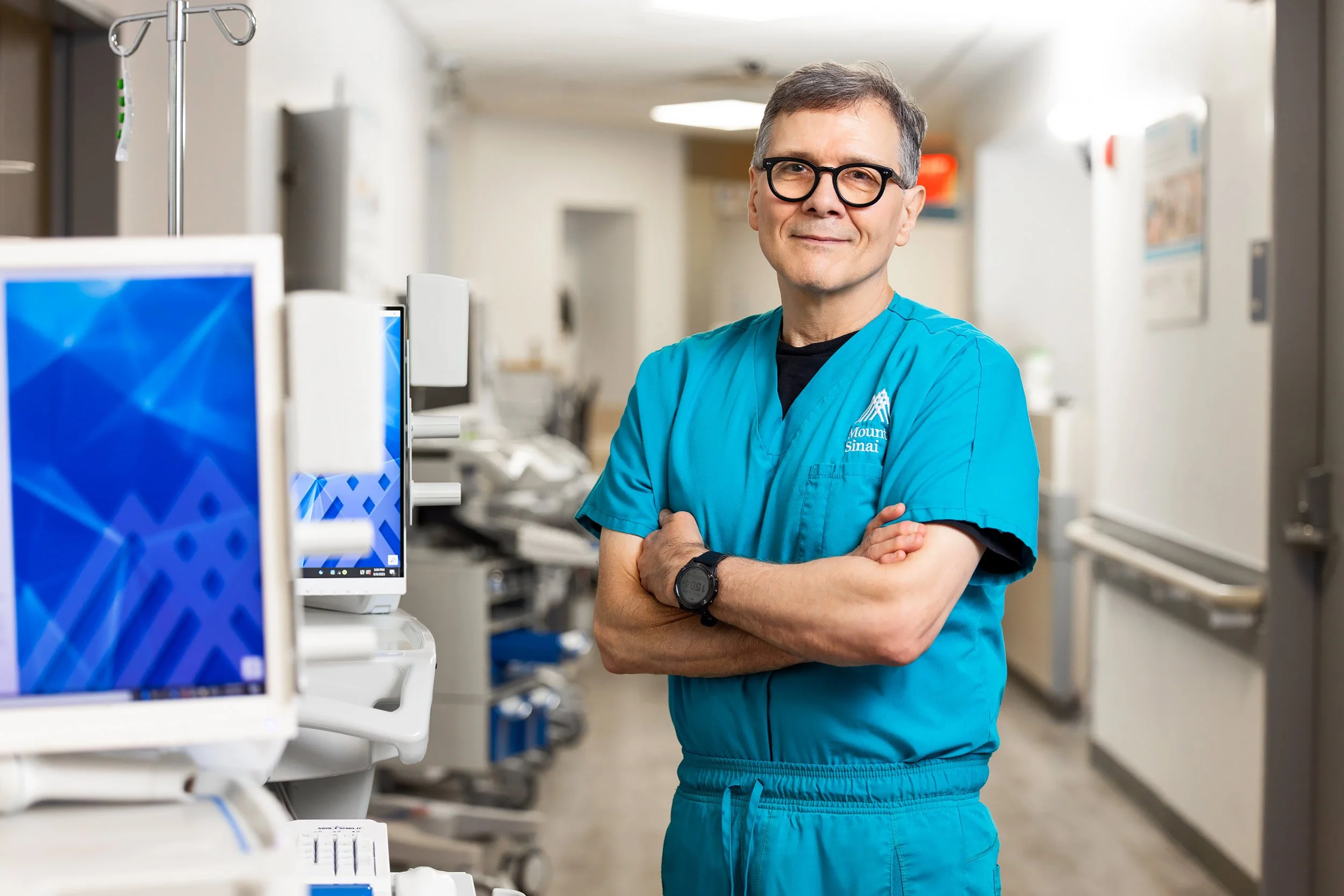 Male doctor in teal scrubs with crossed arms smiling confidently in a hospital corridor.