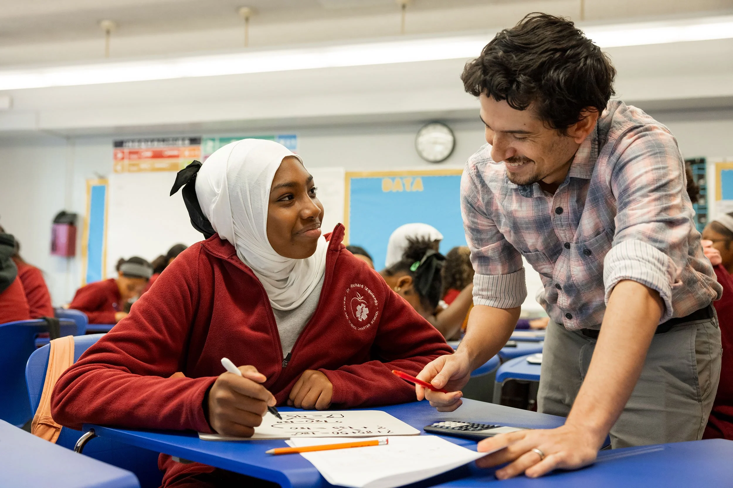 Teacher leaning over desk helping a student with writing assignment in bright classroom.