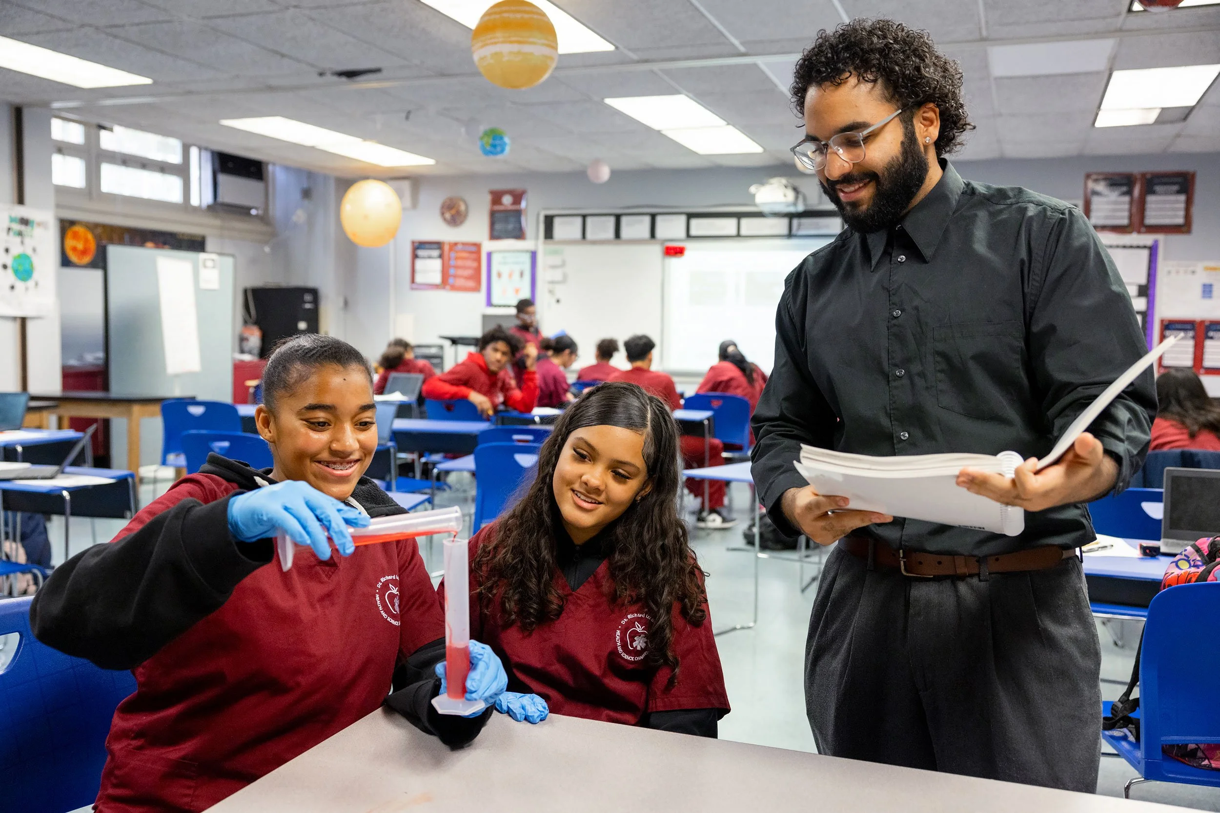 Teacher standing and reading while students sit at a desk using beakers and liquids during science class.
