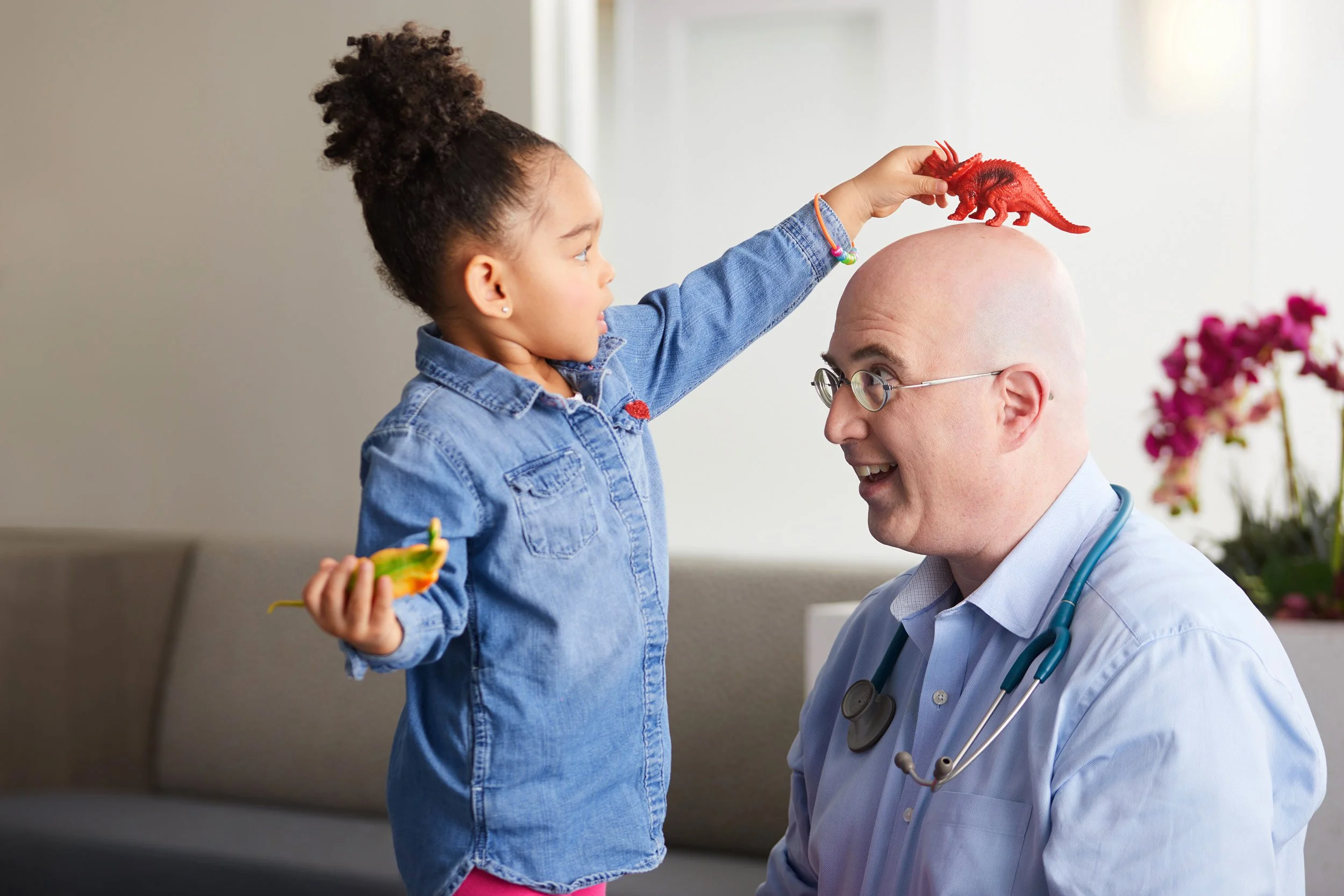 Male doctor with stethoscope and child patient playing together with toy dinosaurs during a pediatric visit.