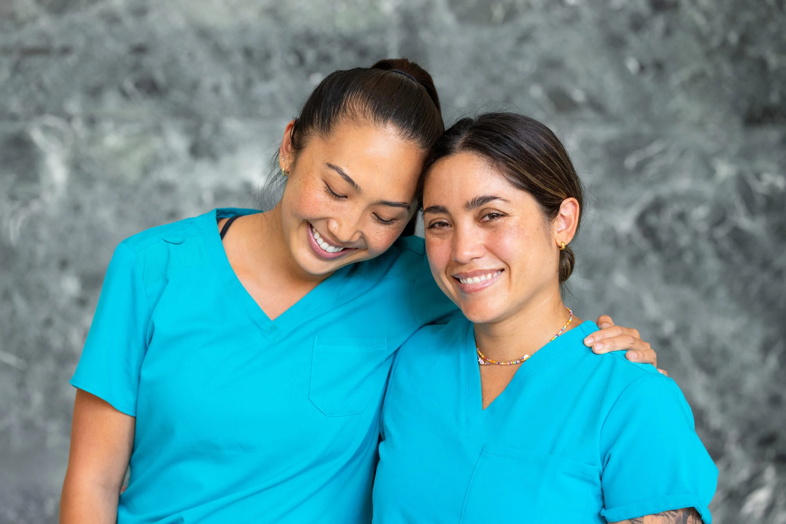 Two nurses smiling together in scrubs against a neutral backdrop.