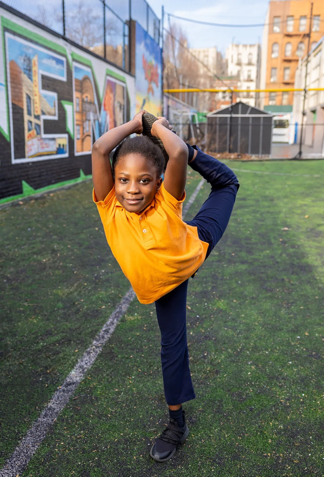 Child in yellow shirt balancing a yoga pose during outdoor playtime on turf field.
