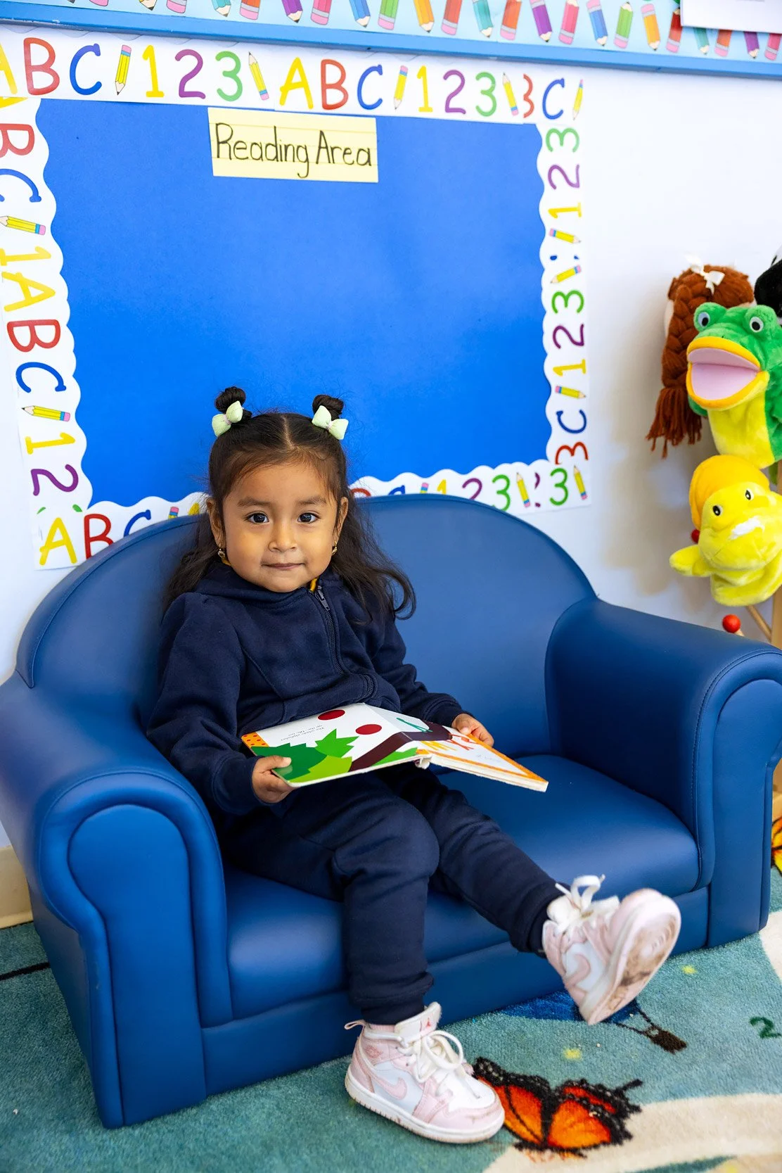 Young child sitting in a blue chair reading a book in a classroom reading corner.
