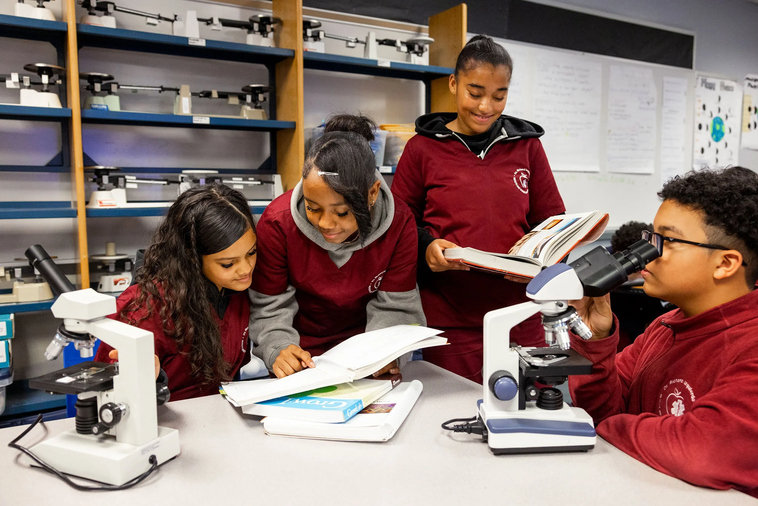 Students collaborating on a science project with books and microscopes spread across a large table.