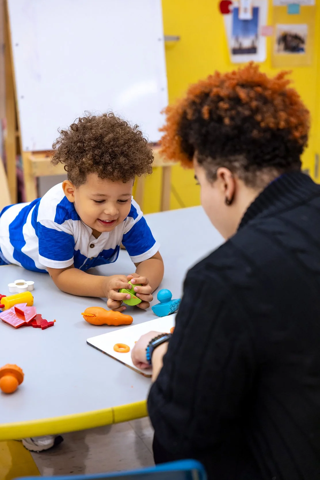 Teacher sitting with young child at a small table, guiding a learning activity with colorful clay.